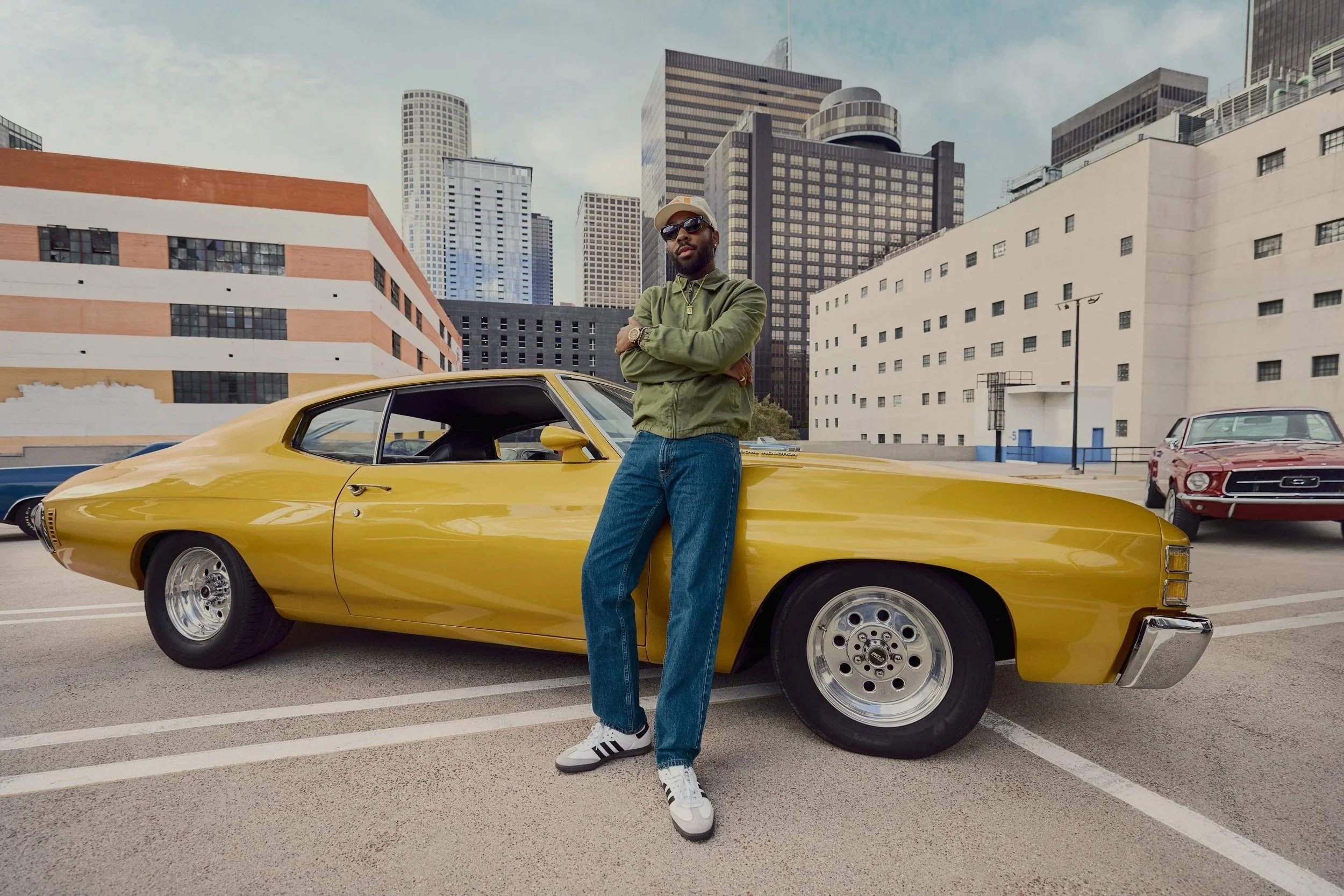 A man stands with arms crossed leaning against a yellow vintage sports car in a parking lot with tall skyscrapers in the background.