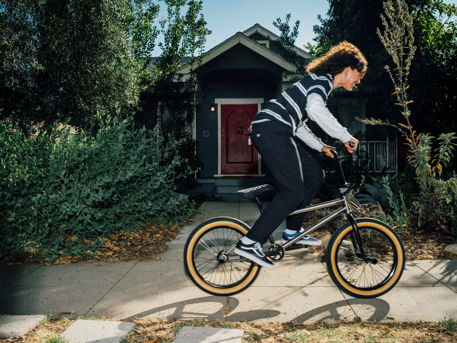 A person with curly hair riding a bicycle on a sidewalk in front of a house with a red door, surrounded by green bushes and trees.