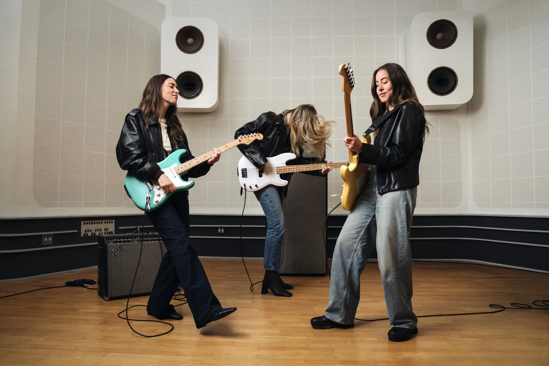 The women of HAIM in black leather jackets playing electric guitars and practicing in a music room with large white speakers on the wall, a small amplifier on the floor, and wooden flooring.