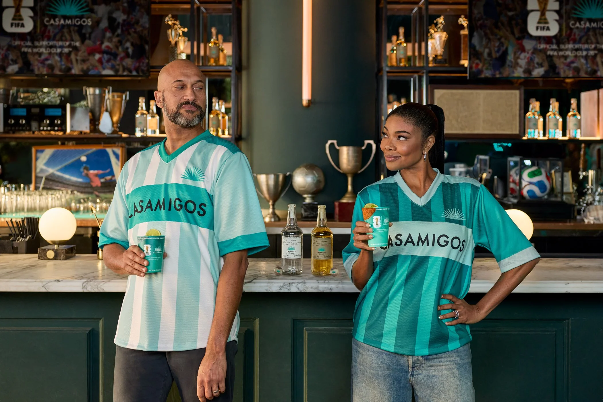 Gabrielle Union and Keegan-Michael Key stand at a bar in a sports-themed setting, both wearing turquoise sports jerseys with 'Casamigos' written on them, holding drinks, with shelves of bottles and trophies in the background.