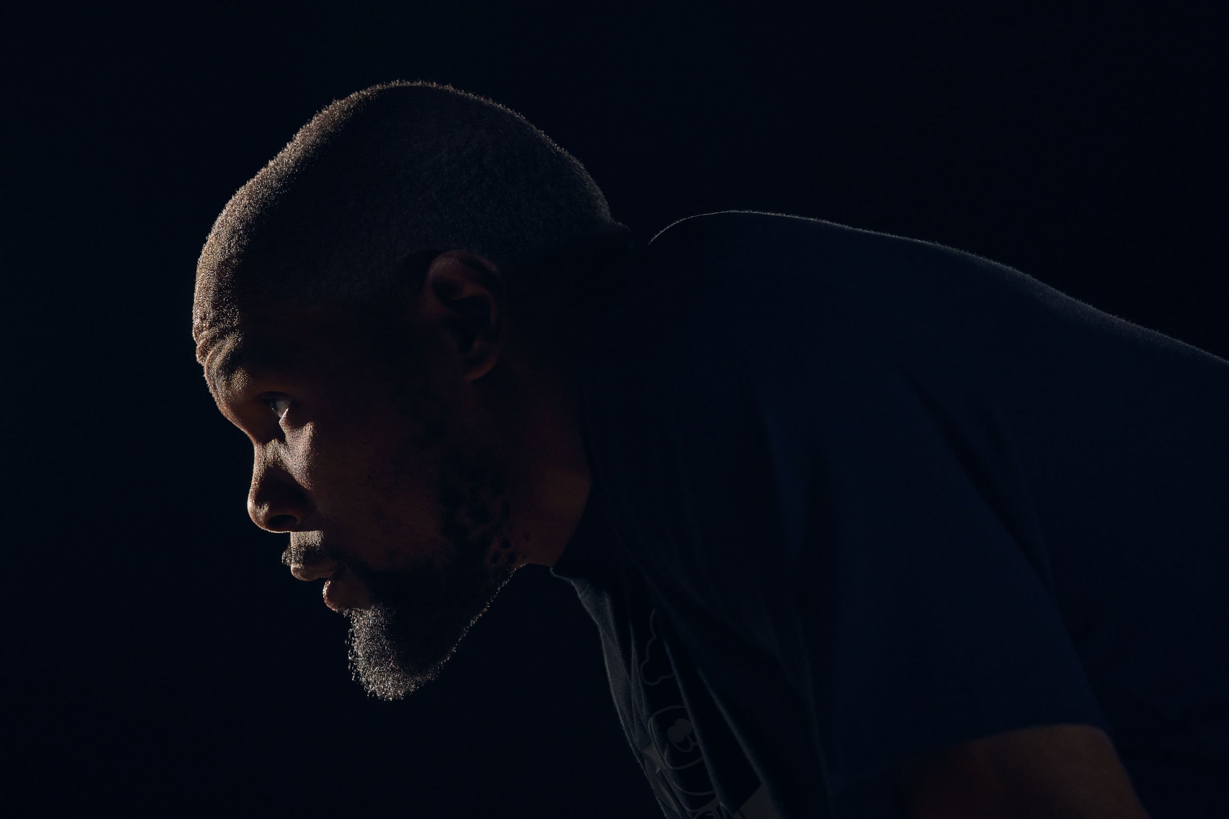 Side profile of Kevin Durant wearing athletic clothing, in a dark setting with minimal lighting highlighting his face, neck, and part of his shoulder.