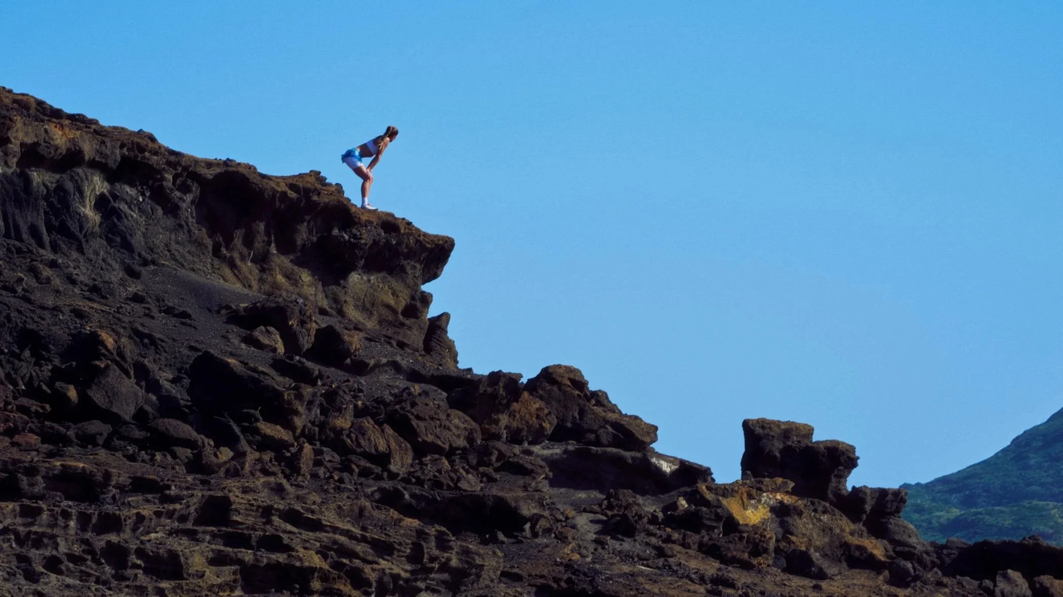 A person wearing a gray shirt and blue shorts standing on a rocky cliff, looking down and leaning forward, with a clear blue sky and mountainous landscape in the background.