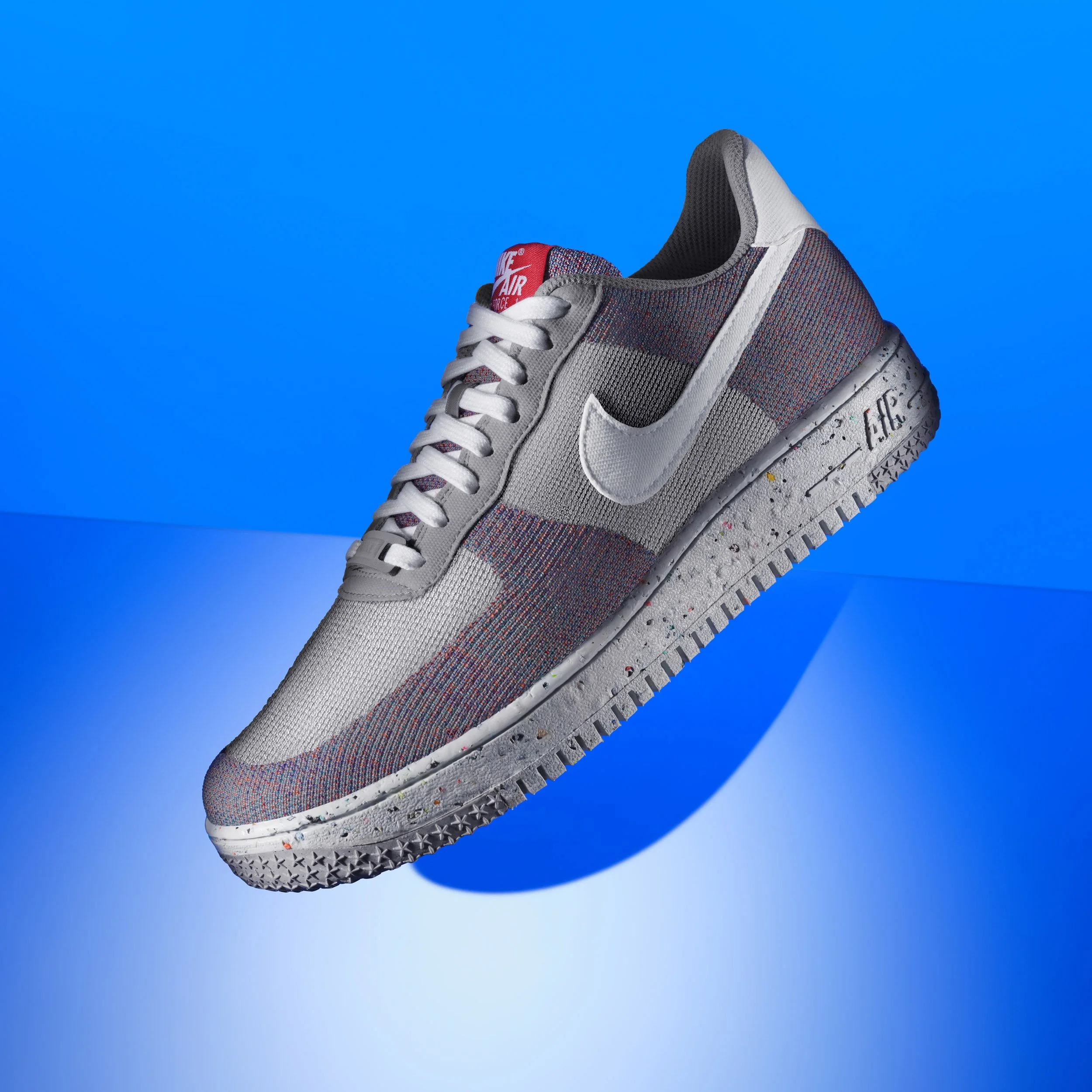 Close-up of a Nike sneaker with a multicolor fabric upper, white laces, a red Nike Air tongue, and a white midsole with colorful speckles, against a blue background.