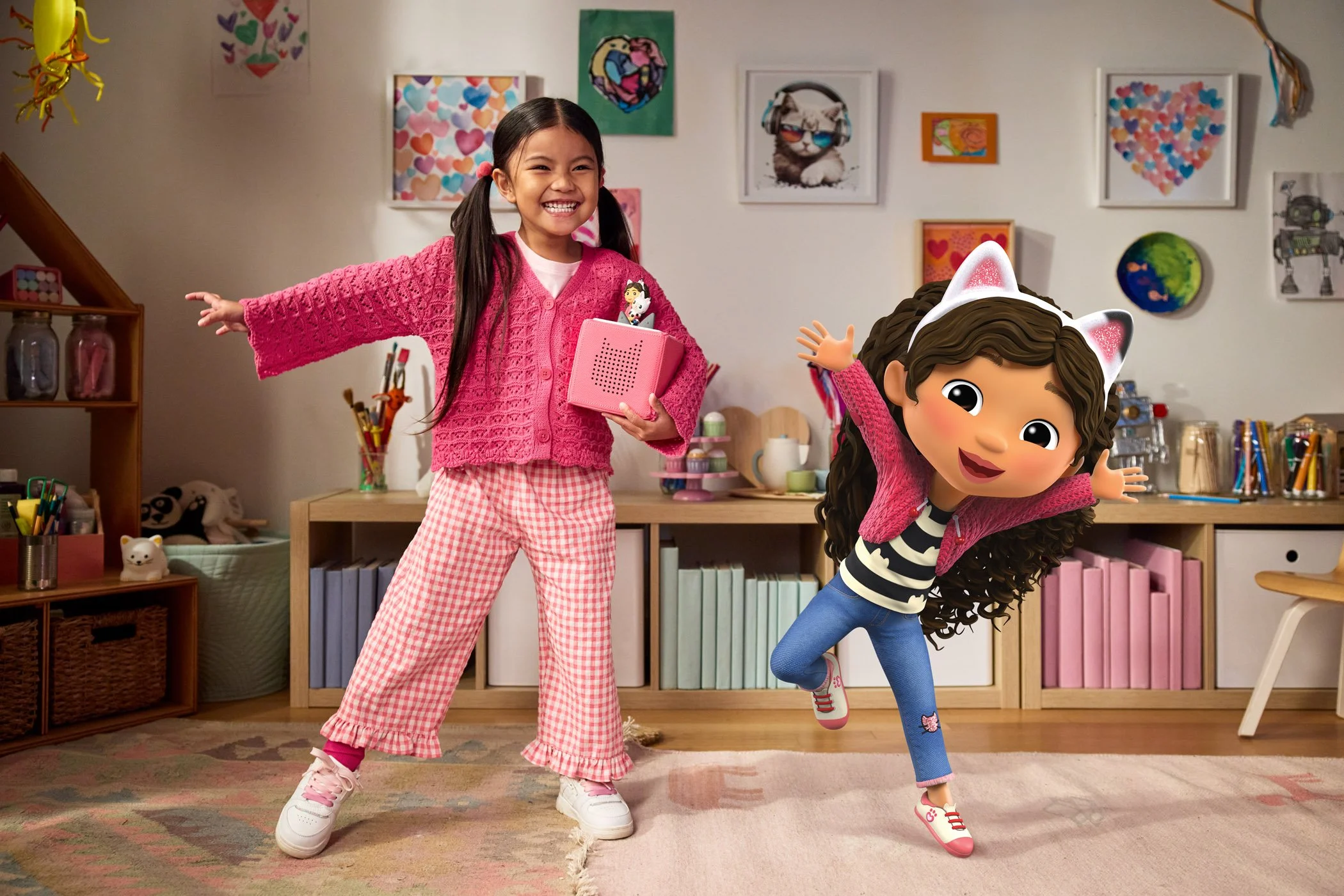 A young girl in a pink sweater and checkered pants smiling and posing with a cartoon character in a room decorated with colorful artwork and toys.