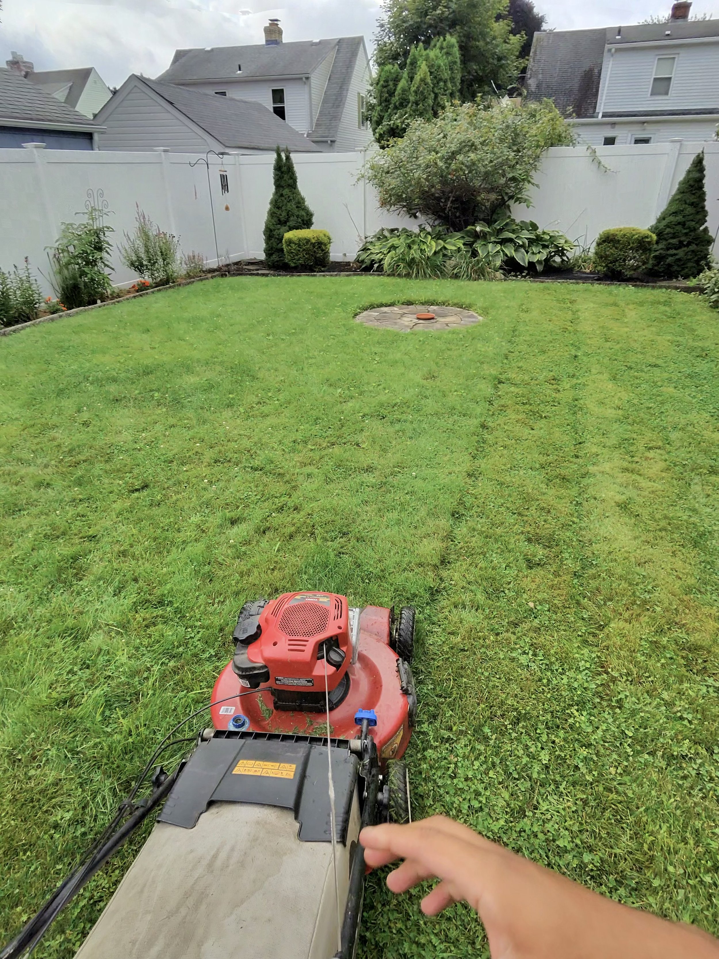 View from a lawnmower operator's perspective, looking towards a green lawn with garden beds and shrubs, enclosed by a white fence, with residential houses in the background under cloudy sky.