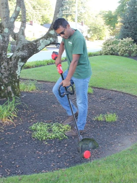 Man using a cordless weed trimmer in a garden area with grass and plants.