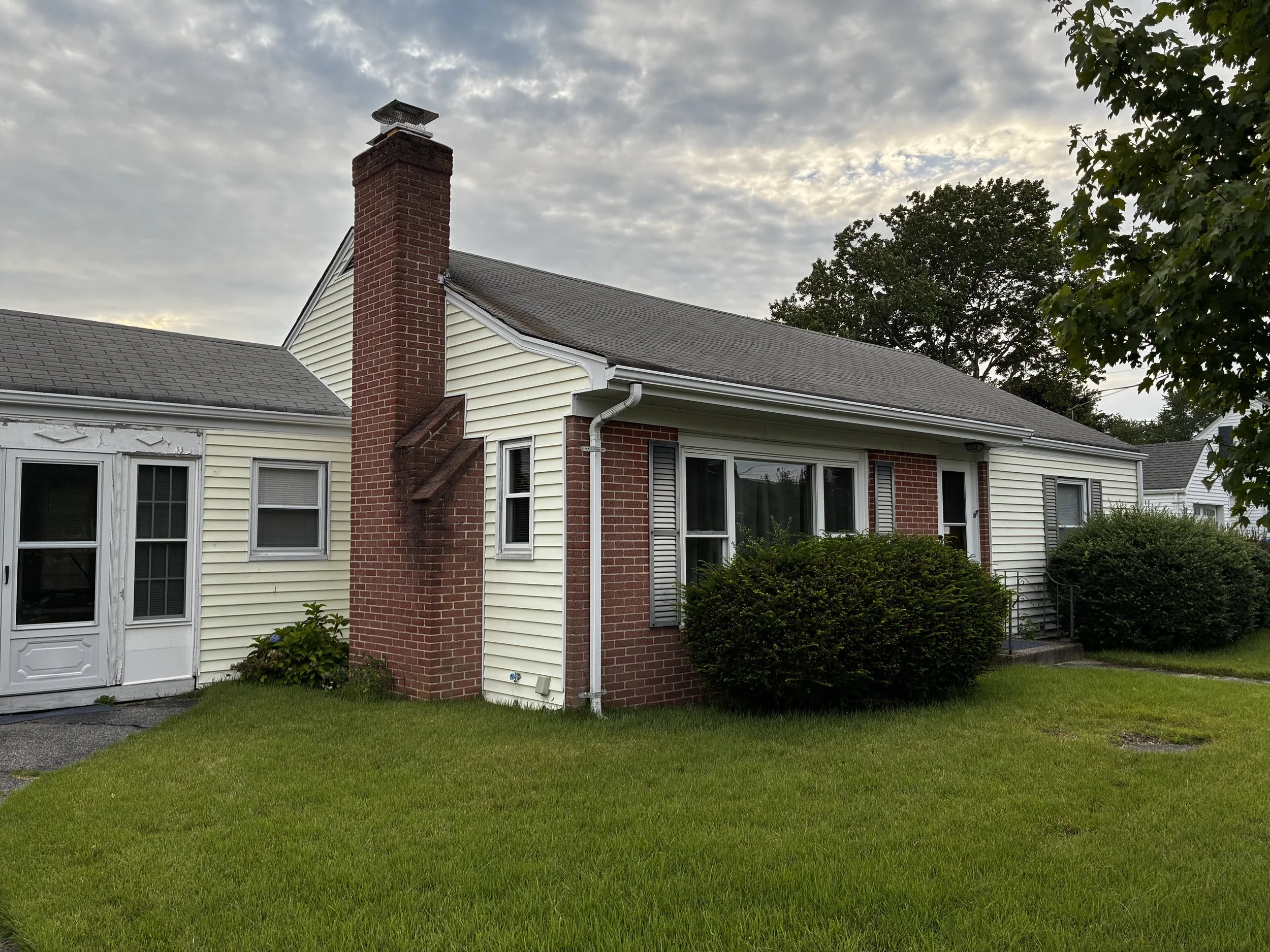 A house with a brick chimney, yellow siding, white trim, and a slate roof, with a grassy lawn and trees in the background under a cloudy sky.