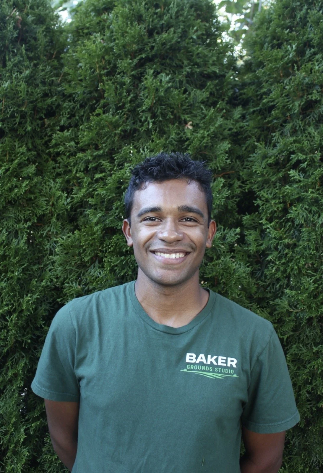 Smiling young man outdoors in front of green bushes, wearing a green t-shirt with 'Baker Grounds Studio' logo.