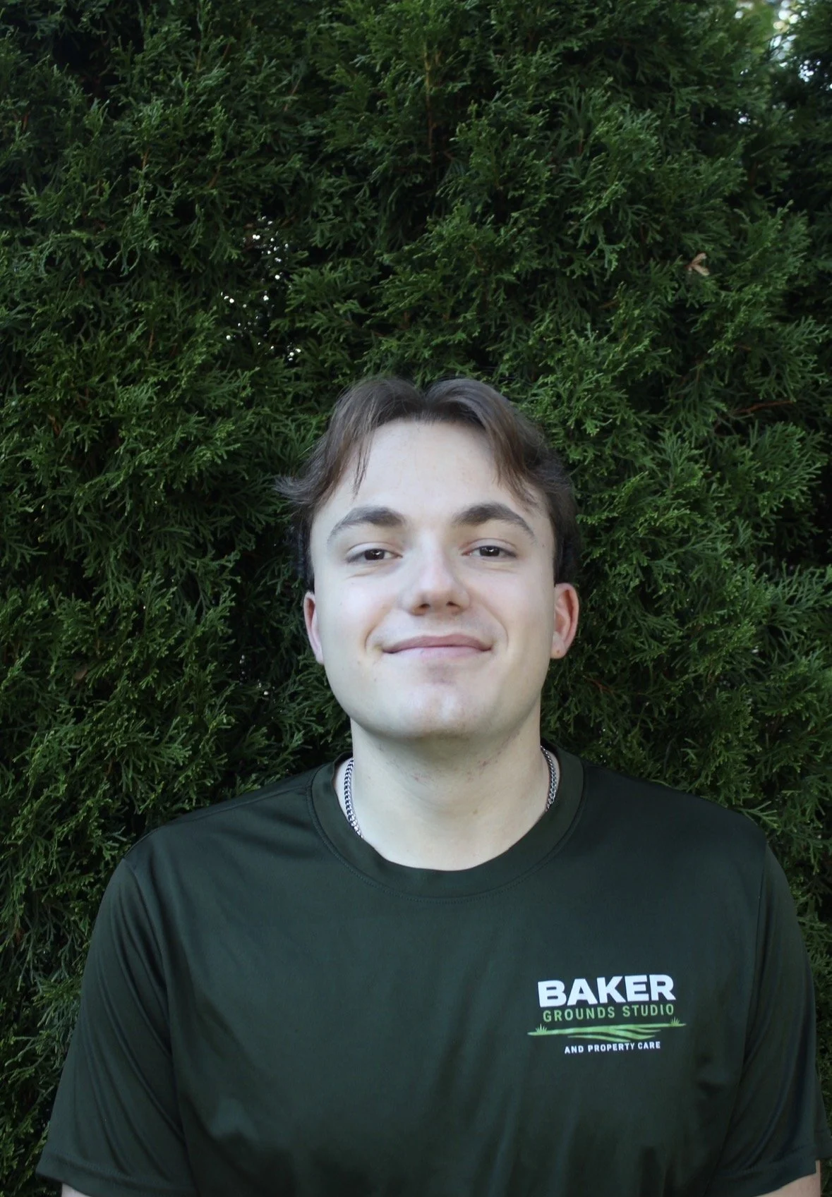 A young man with light skin, short brown hair, and a slight smile standing in front of a dense green bush. He is wearing a black T-shirt with a logo that reads 'BAKER GROUNDS STUDIO AND PROPERTY CARE.'
