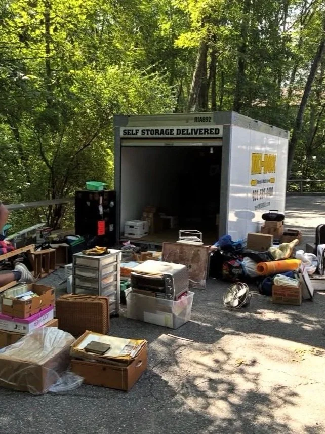 A moving truck parked on the side of a road with its back open, surrounded by various boxes, furniture, and household items scattered on the ground. The area is shaded by trees.