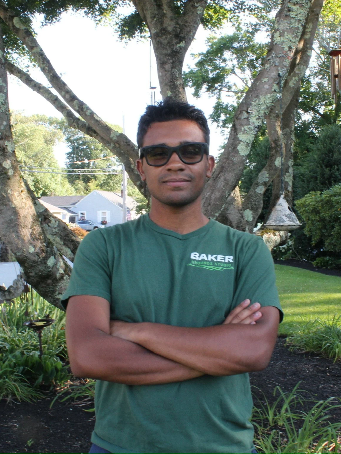 A young man wearing sunglasses and a green T-shirt with a logo stands outdoors in front of a large tree with a swing. His arms are crossed, and he has short black hair and a slight smile.