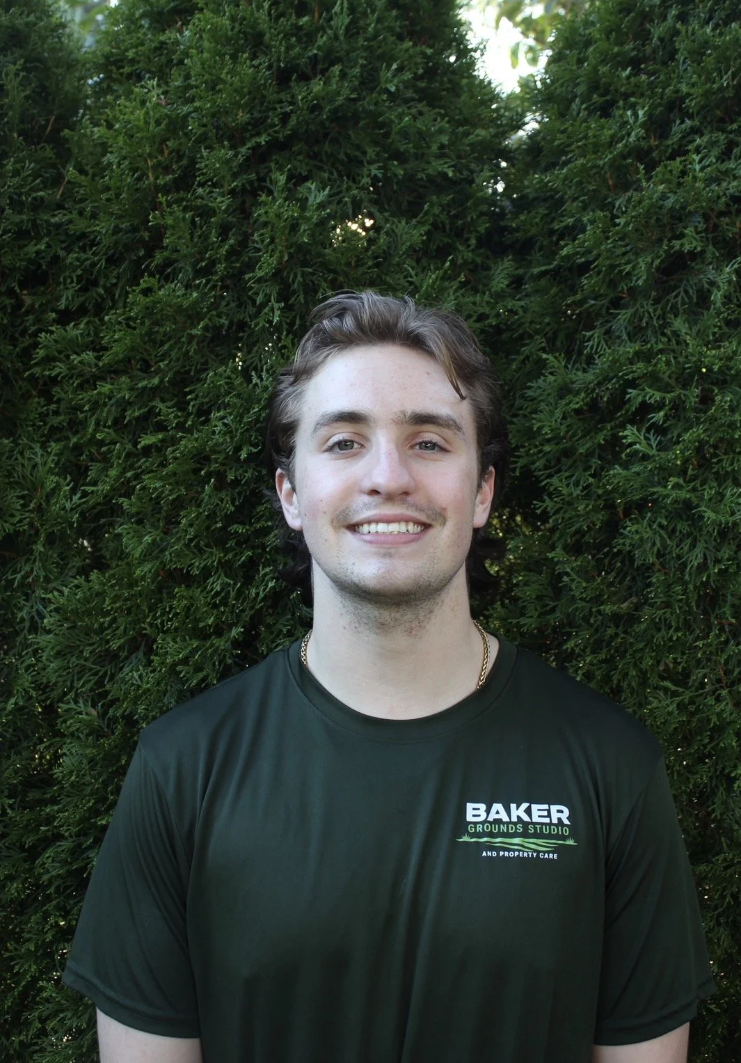 A young man with light brown hair and a smile, wearing a black T-shirt that reads 'Baker Grounds Studio and Property Care', standing in front of green bushes.