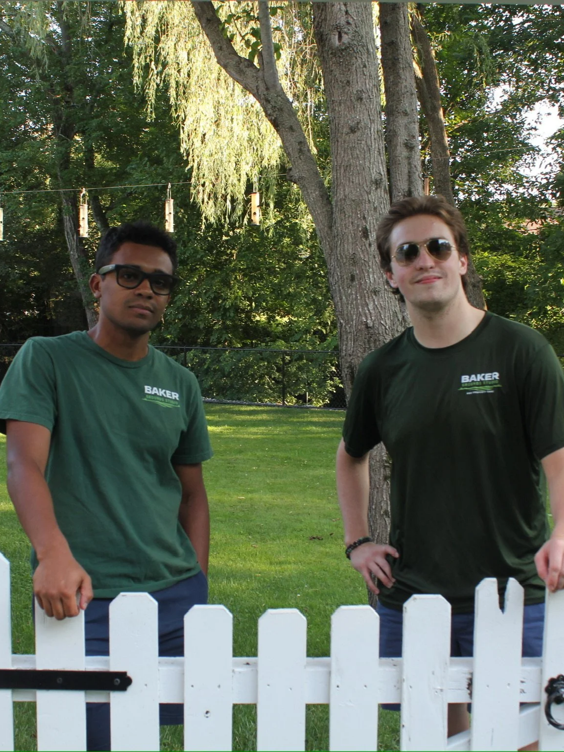 Two young men standing outdoors behind a white picket fence, with large trees and birdhouses in the background. Both are wearing green shirts with the word 'Baker' on them, one with sunglasses and the other with glasses.