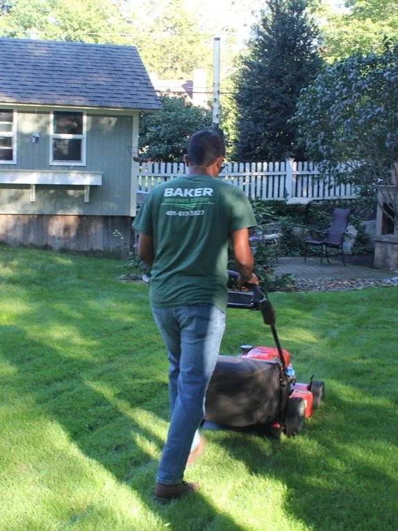 Person pushing a red lawn mower in a backyard with green grass, trees, and a gray house with a white fence in the background.