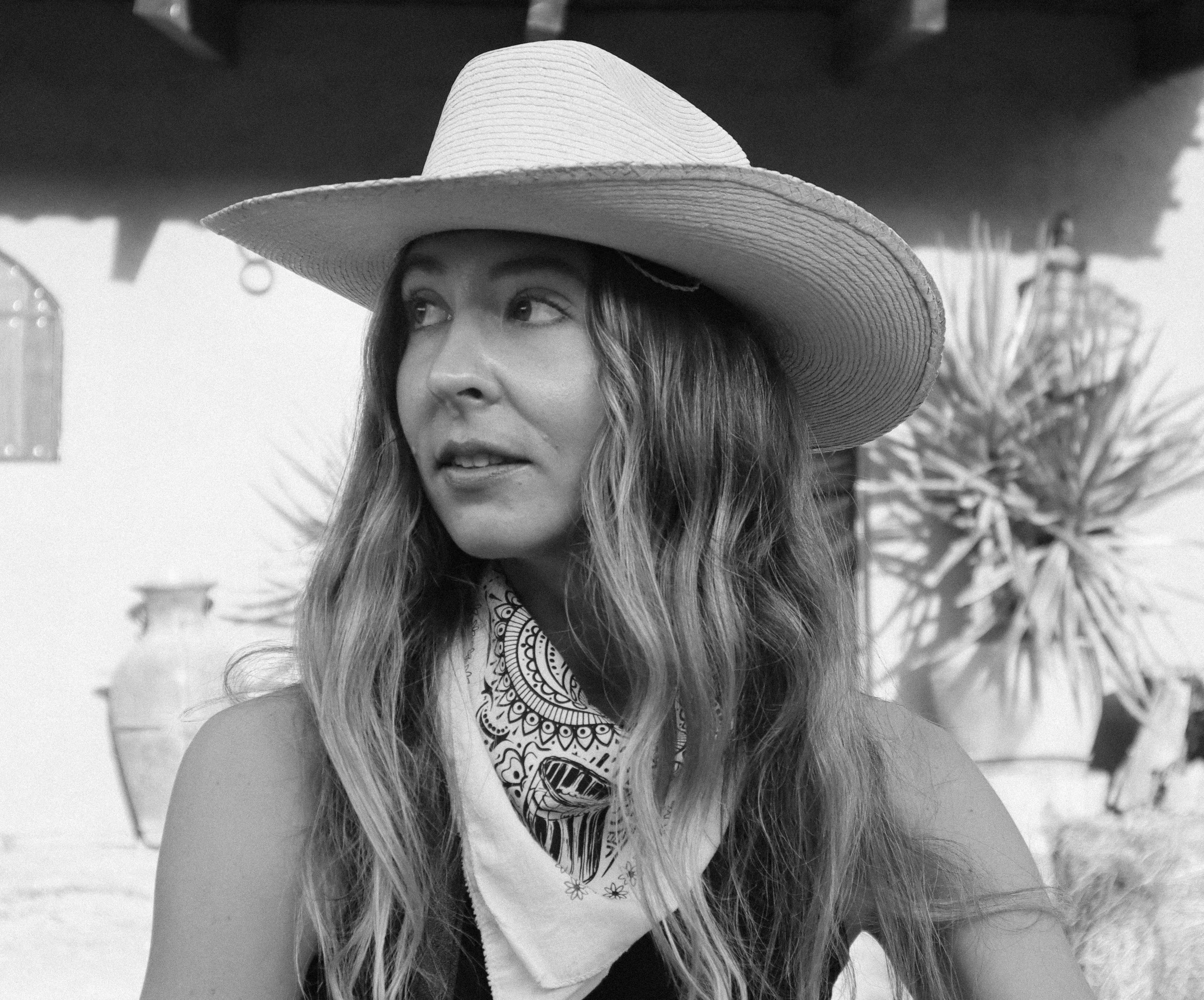A woman with long hair wearing a large sunhat and a bandana around her neck, standing outdoors with palm trees and a building in the background.