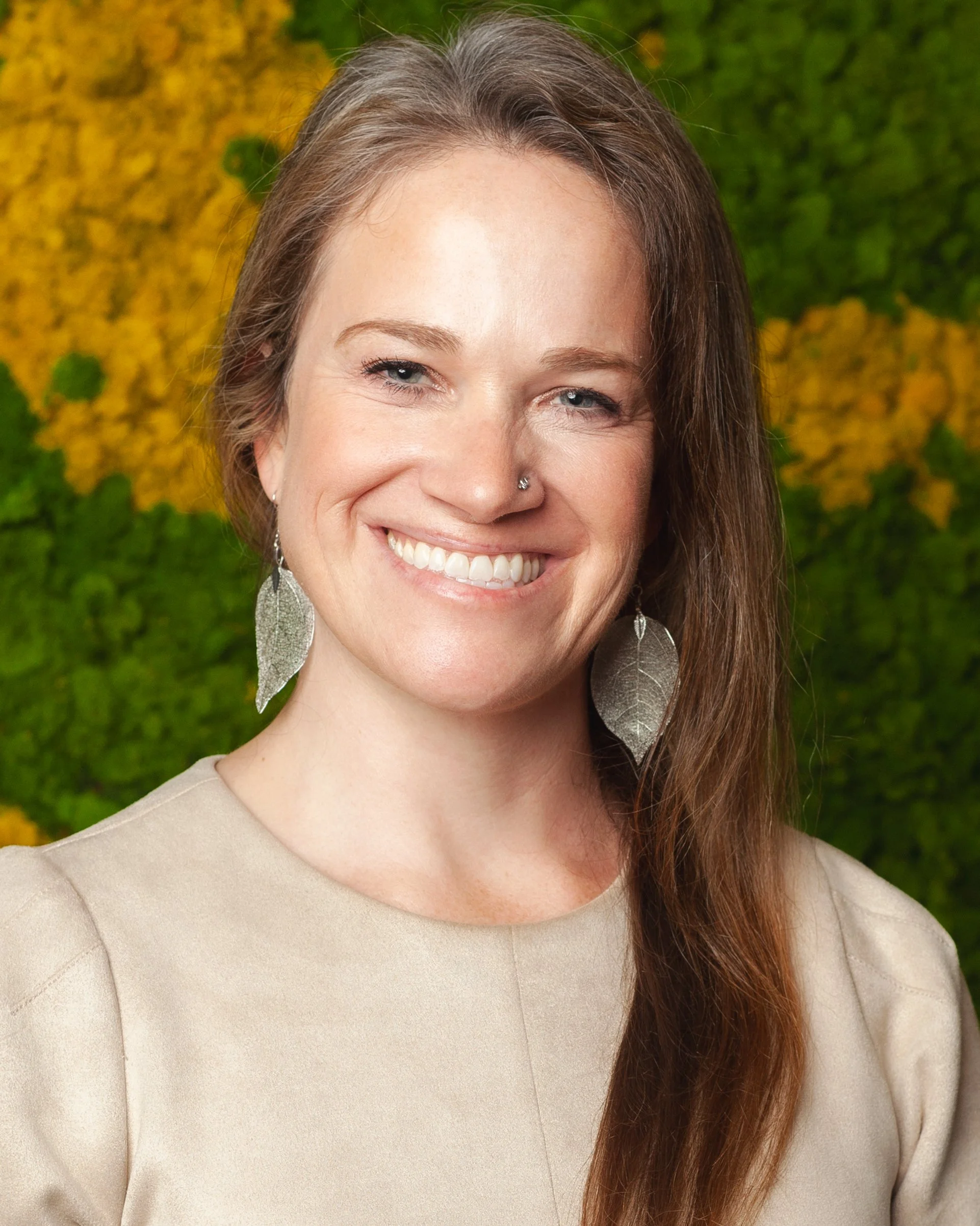 A woman smiling outdoors with greenery and yellow flowers in the background. She has long brown hair, blue eyes, a nose piercing, and is wearing a beige top and large leaf-shaped earrings.