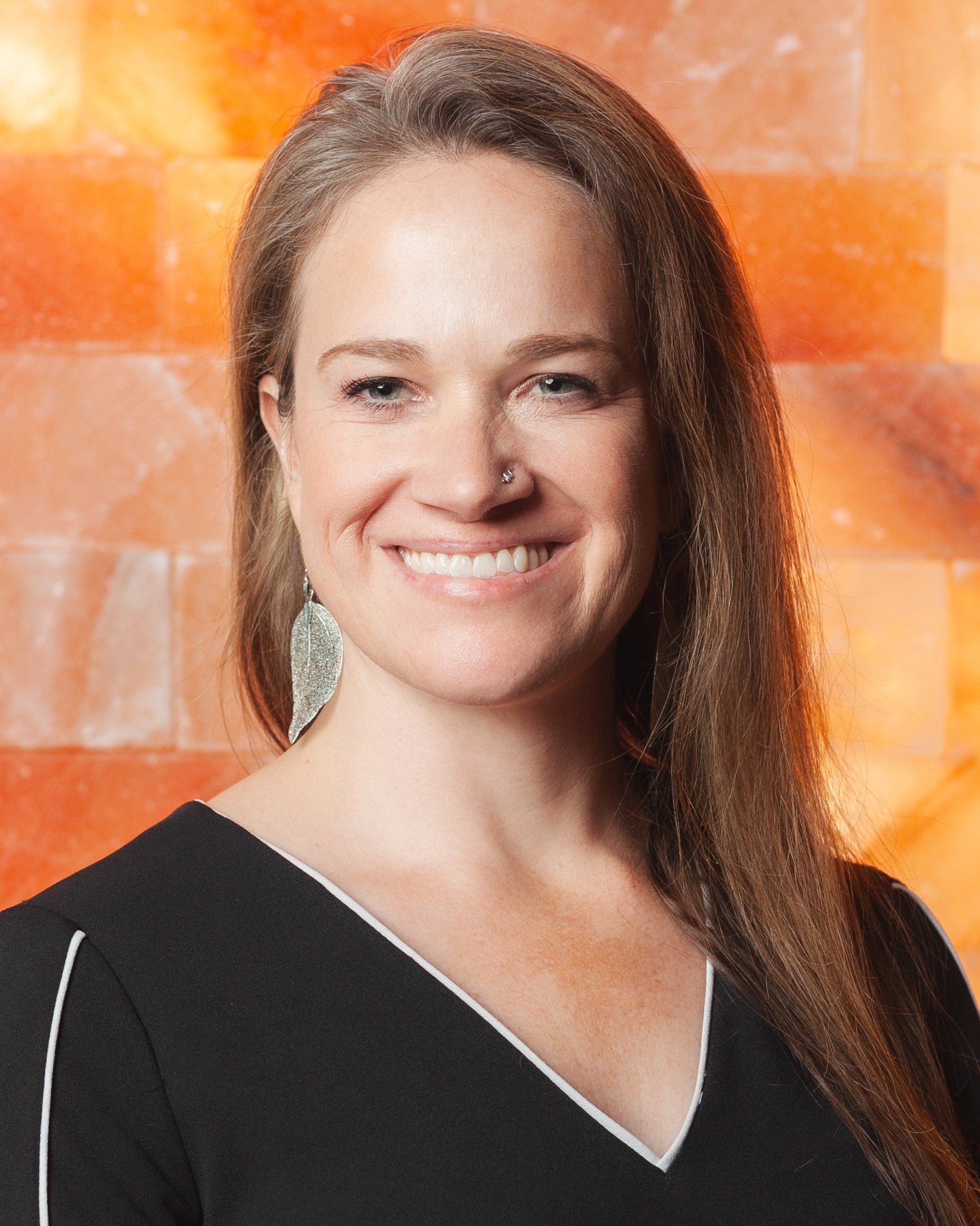 A smiling woman with long brown hair, wearing a black top with white trim, earrings, and a nose ring, standing in front of an orange brick wall.