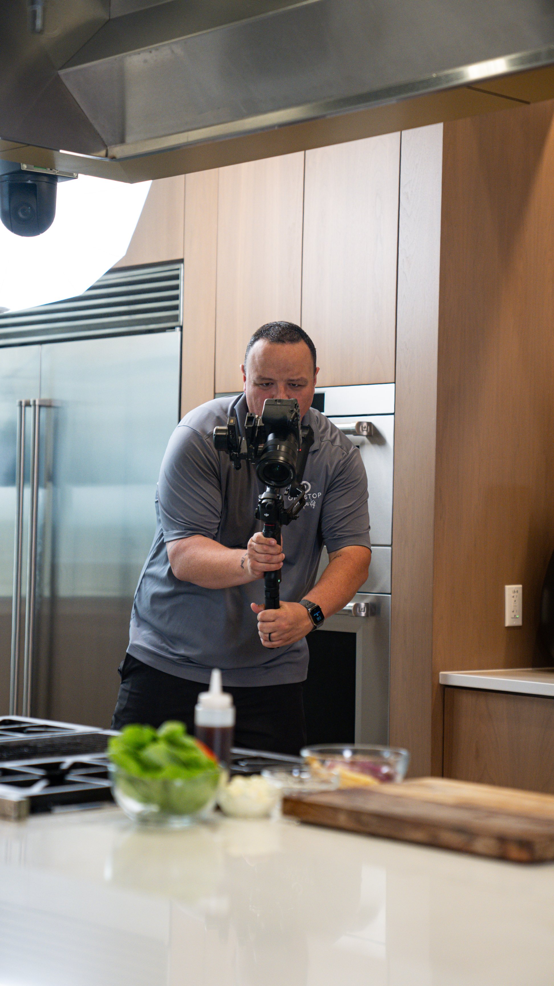 A man in a gray shirt operating a camera in a kitchen with wooden cabinets and stainless steel appliances, with cooking ingredients and utensils on the counter in the foreground.