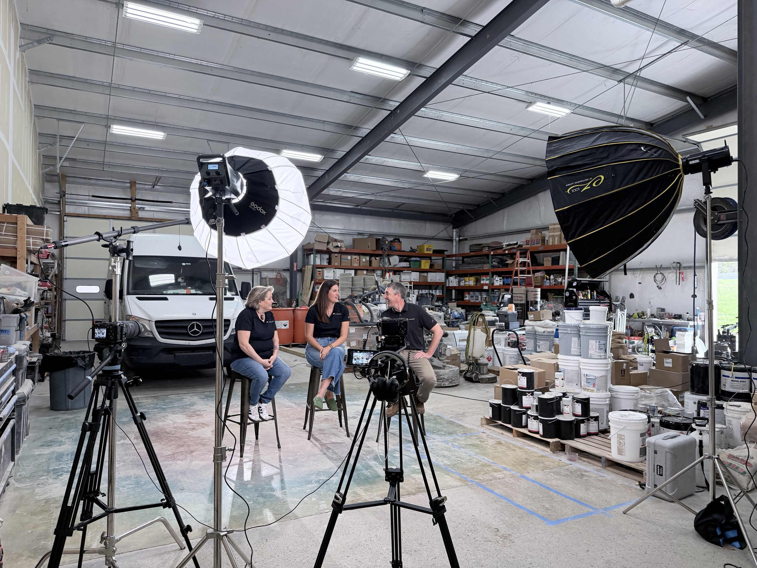 Inside a warehouse converted into a photo studio, three people are sitting on chairs in front of camera equipment and lights. The warehouse contains shelves, boxes, and various supplies, with a white van parked inside. Professional photography lighting equipment is set up, including large softbox lights and a backdrop. The ceiling is high with exposed beams and lighting fixtures.