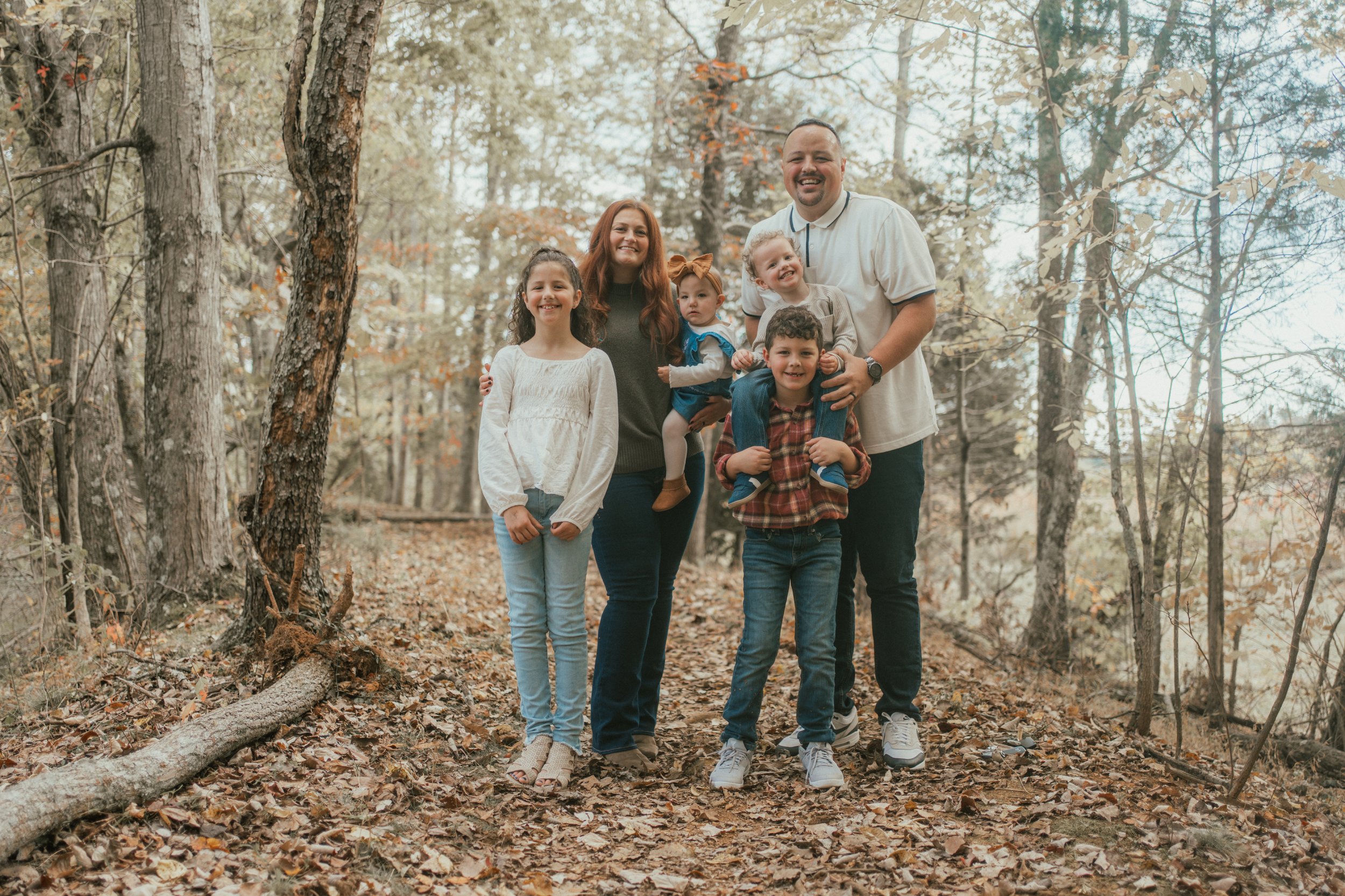 A family of six smiling and posing for a photo in a wooded area during fall. The group includes two adults and four children, standing close together among trees and fallen leaves.