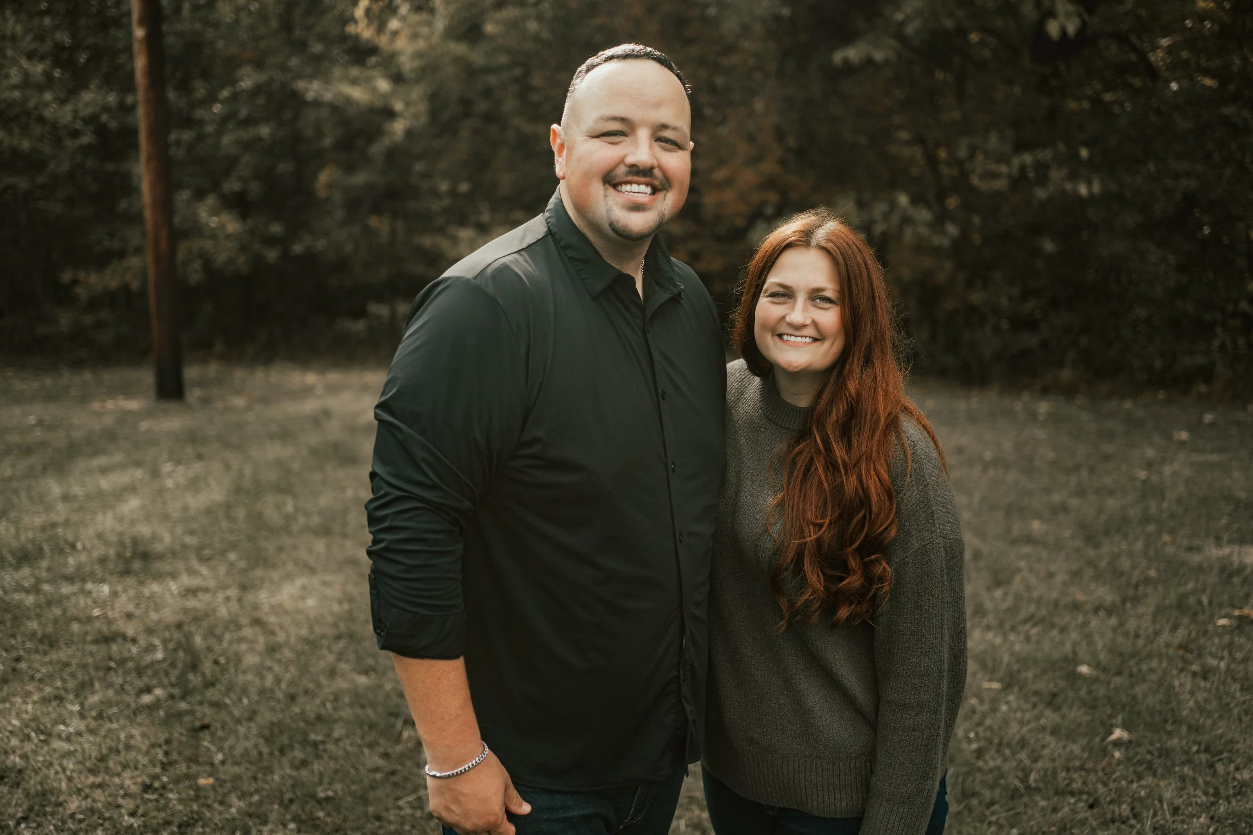 A man and a woman smiling outdoors, standing close together with wooded background.