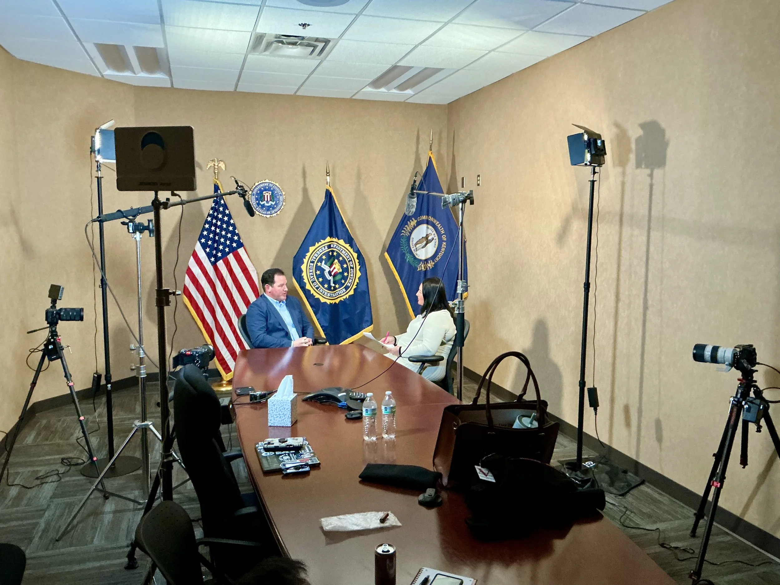 A conference room setup with a long table, chairs, and various recording and lighting equipment. Two people are seated at the table, engaged in conversation, with flags of the United States and the FBI behind them.