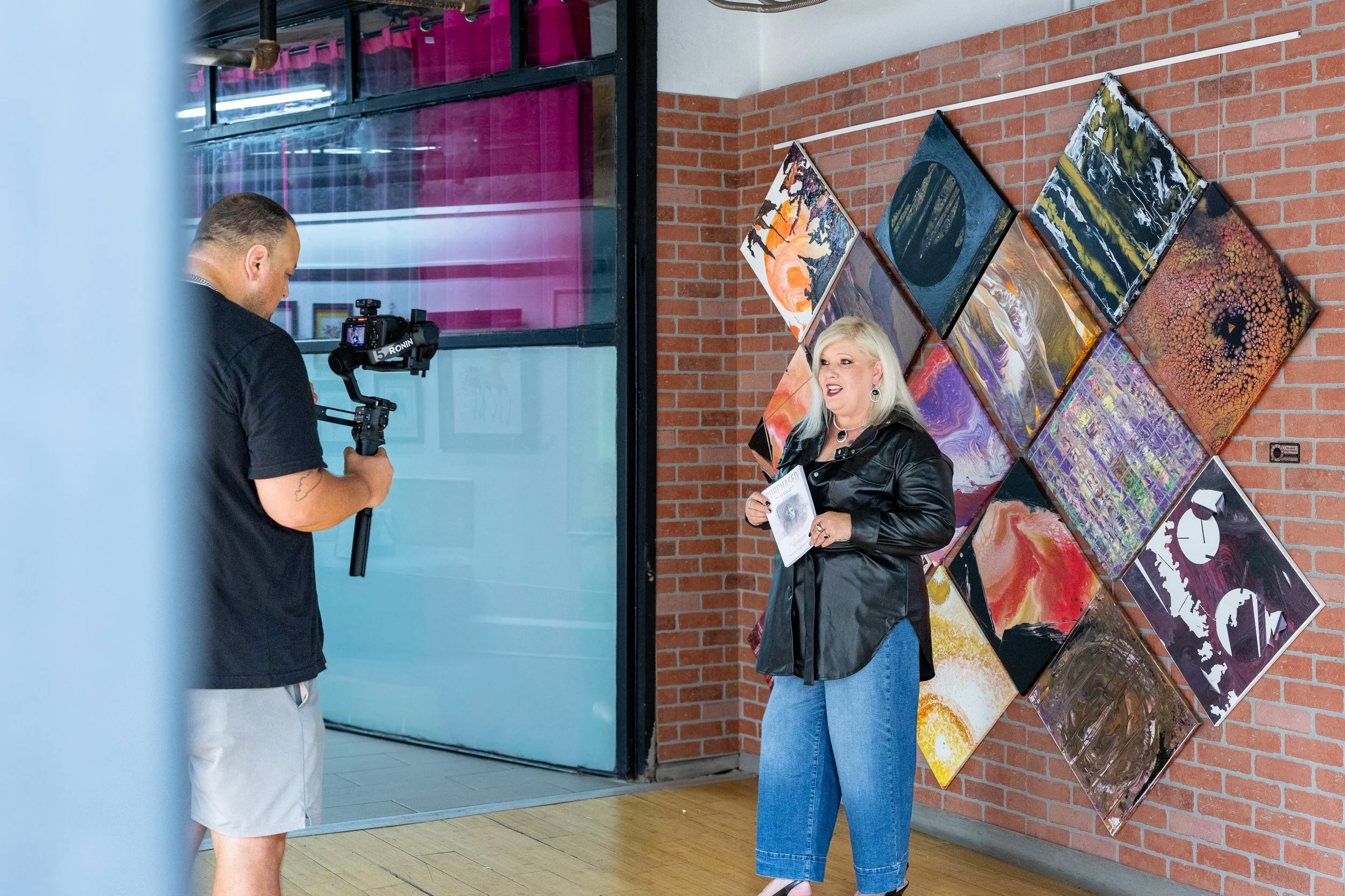 A woman with platinum blonde hair and a black leather jacket is speaking to a man while holding a brochure, in an art gallery. The man is holding a camera on a stabilizer. There are colorful abstract paintings on a brick wall behind her.