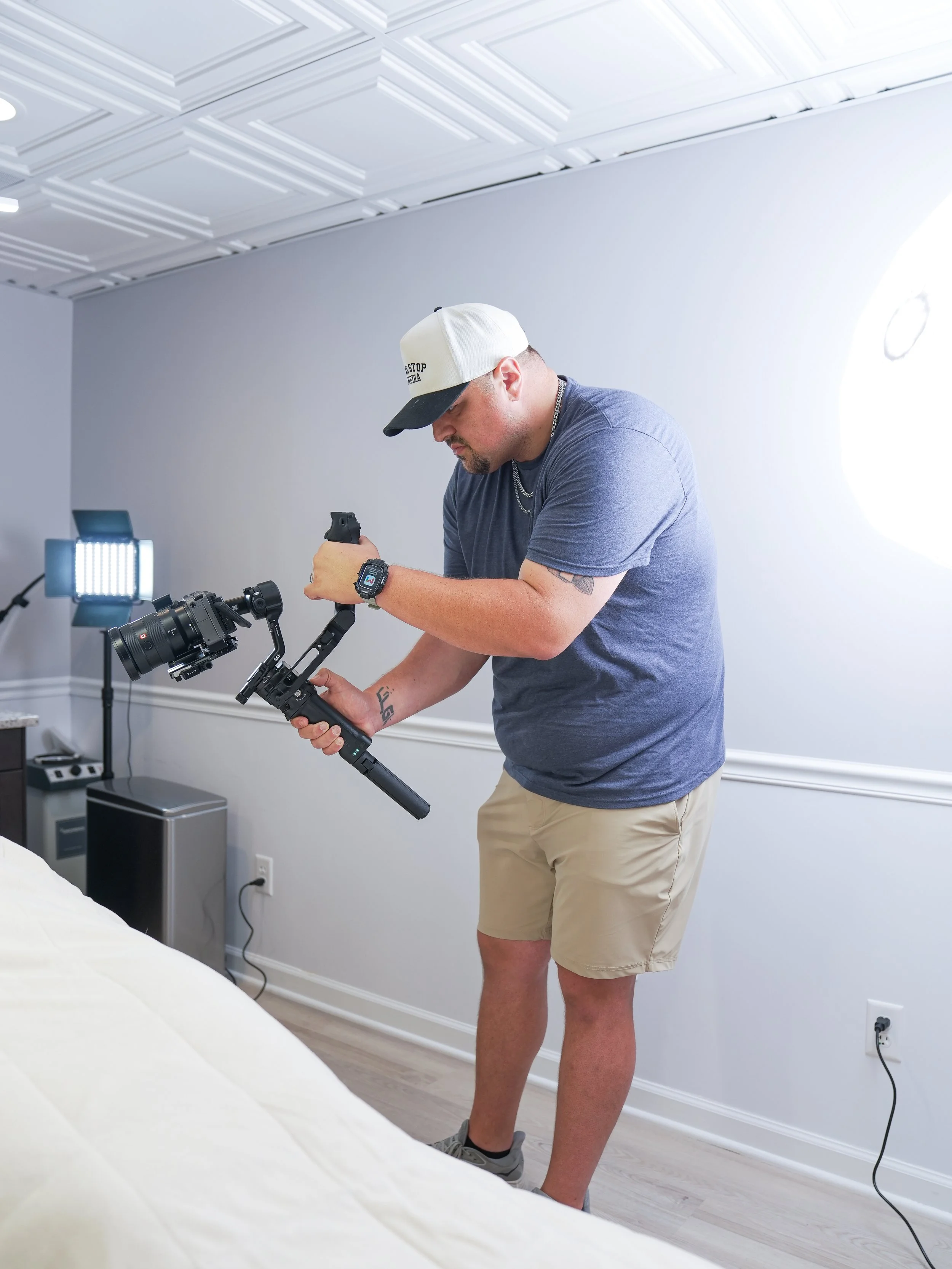 A man wearing a baseball cap, gray T-shirt, khaki shorts, and gray sneakers is holding a camera stabilizer while standing in a room with a white wall, ceiling, and a window with sunlight. He is looking down at the equipment.