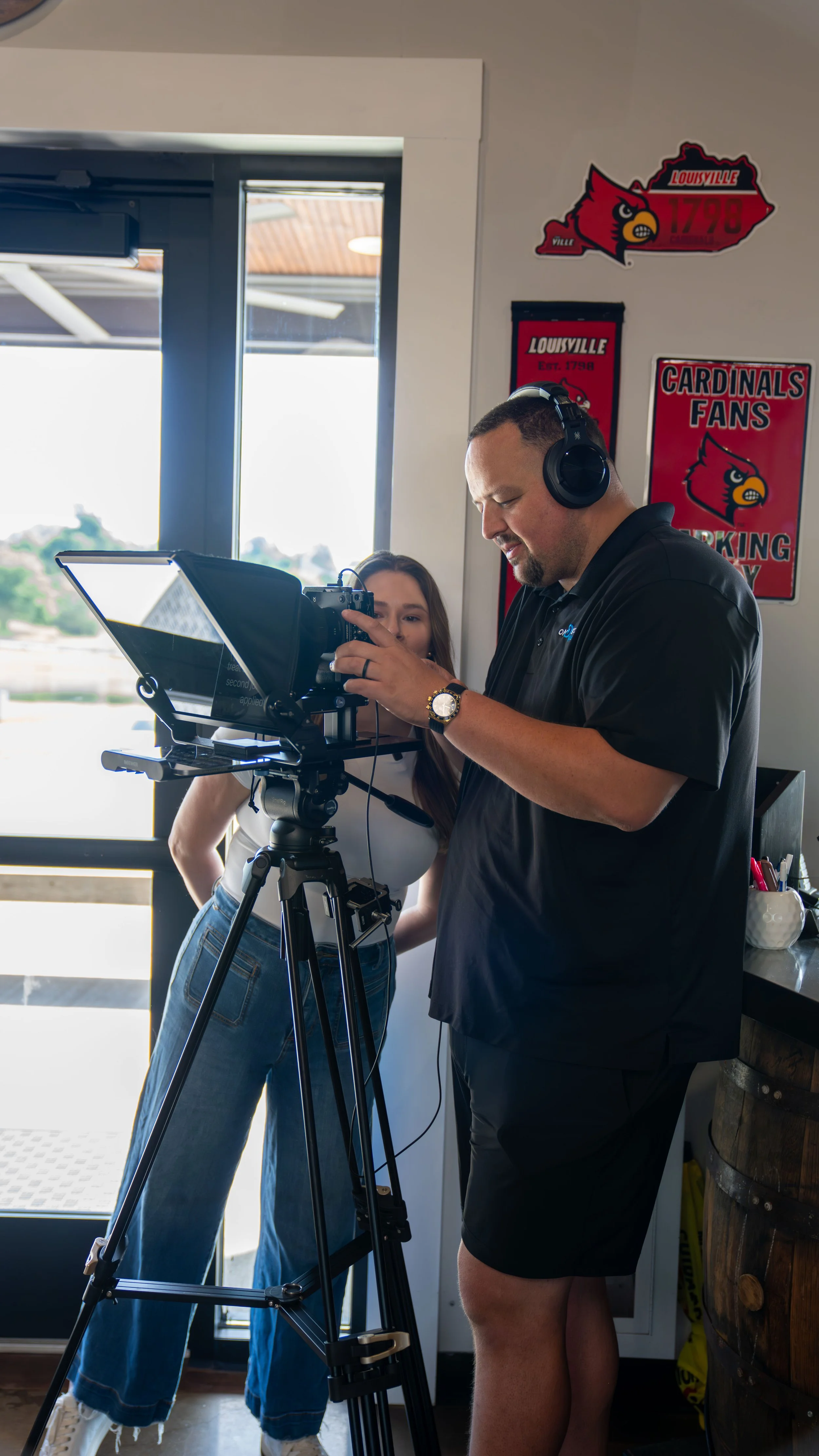 Two people, a man and a woman, are indoors at a camera setup. The man is wearing headphones, a black polo shirt, and shorts, and is looking at a camera mounted on a tripod. The woman, wearing a white top and high-waisted jeans, is standing behind him, partially visible, smiling and looking at the camera. The background has Louisville Cardinals sports memorabilia, including signs and banners.