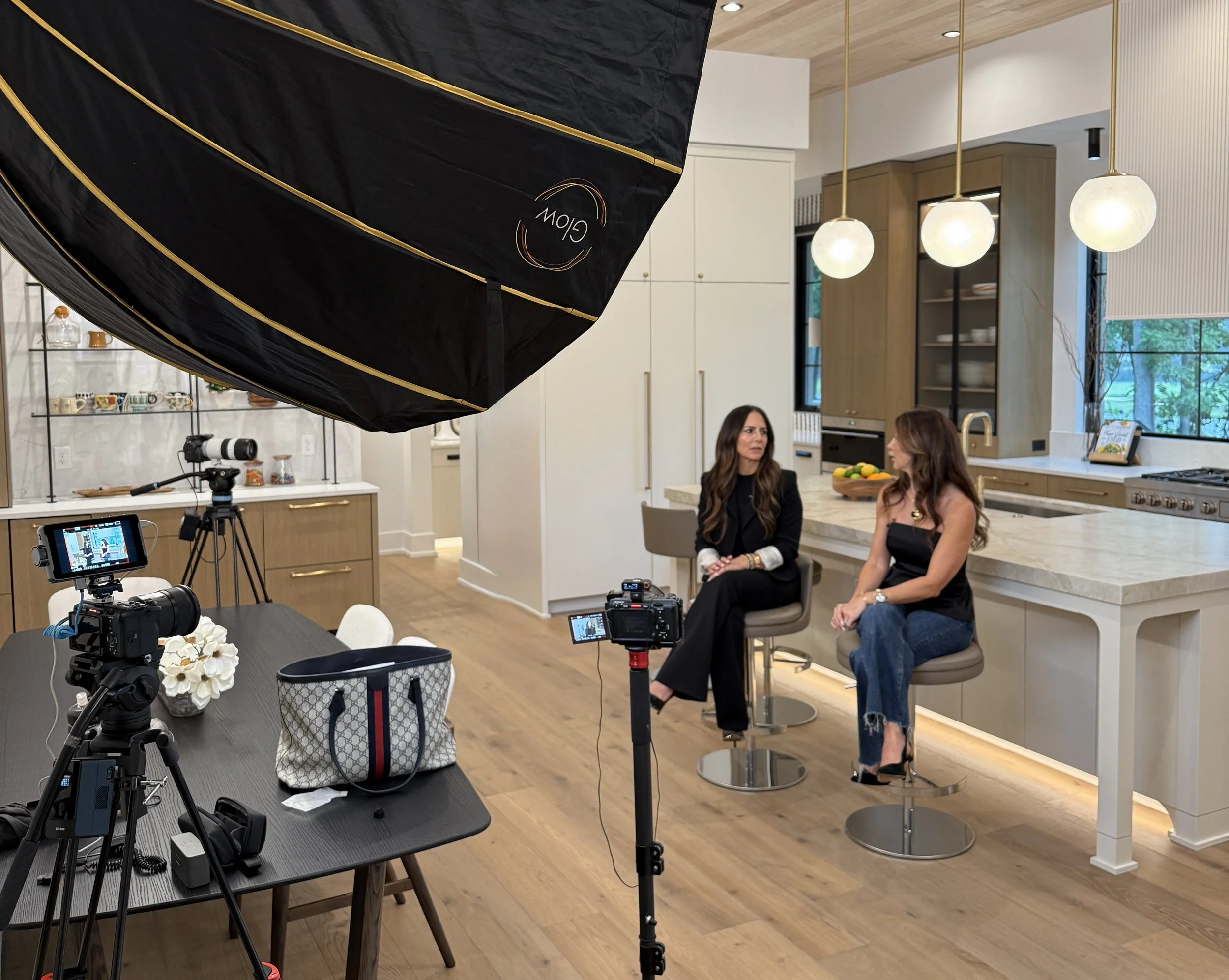 Two women sitting in kitchen, being filmed with camera and lighting equipment in front of them, kitchen with wood cabinets, white walls, hanging pendant lights, and large window showing outdoor greenery.