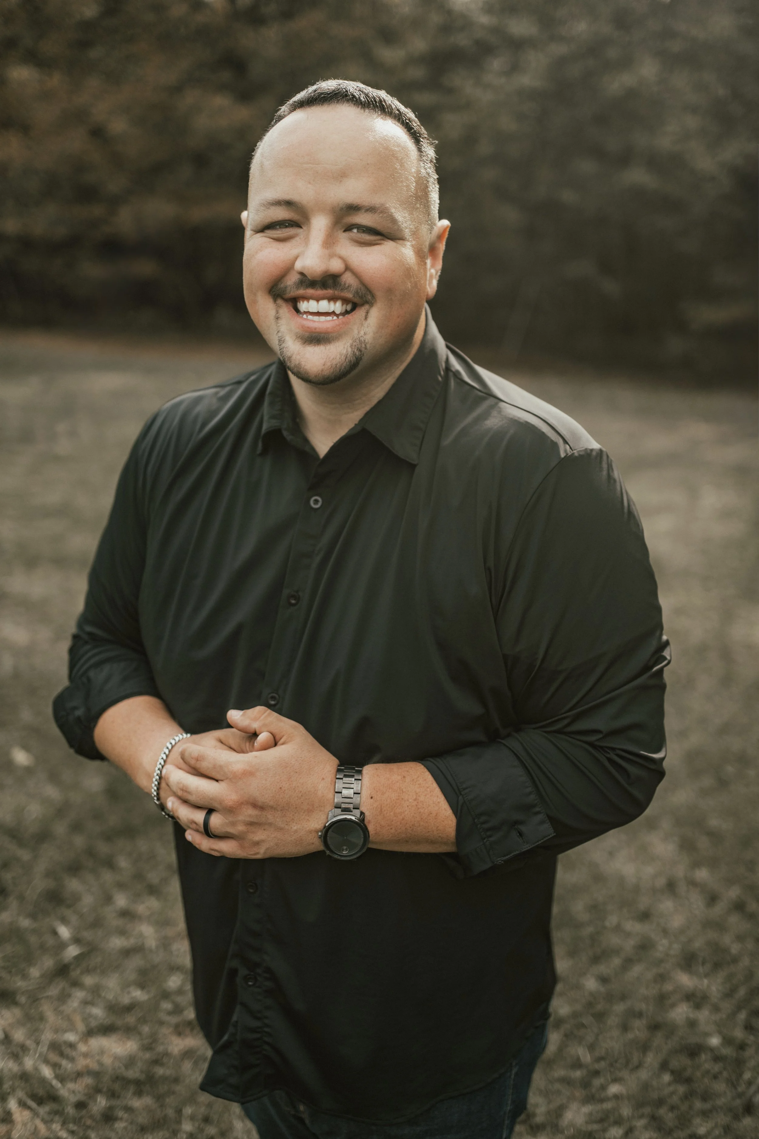 A smiling man with short dark hair and a goatee, wearing a black button-up shirt, standing outdoors in a natural setting with trees and grass in the background.