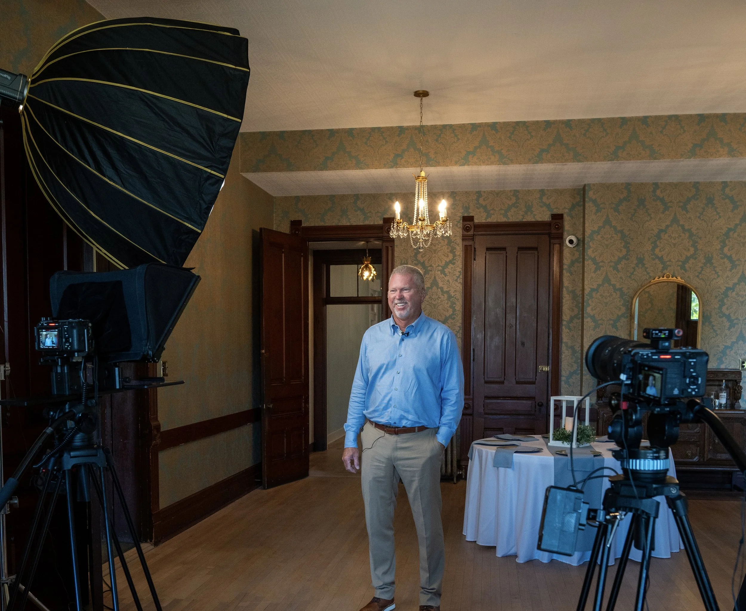 A man stands in a room with ornate wallpaper, wooden doors, and a chandelier, smiling for a photo while cameras and lighting equipment are set up around him.