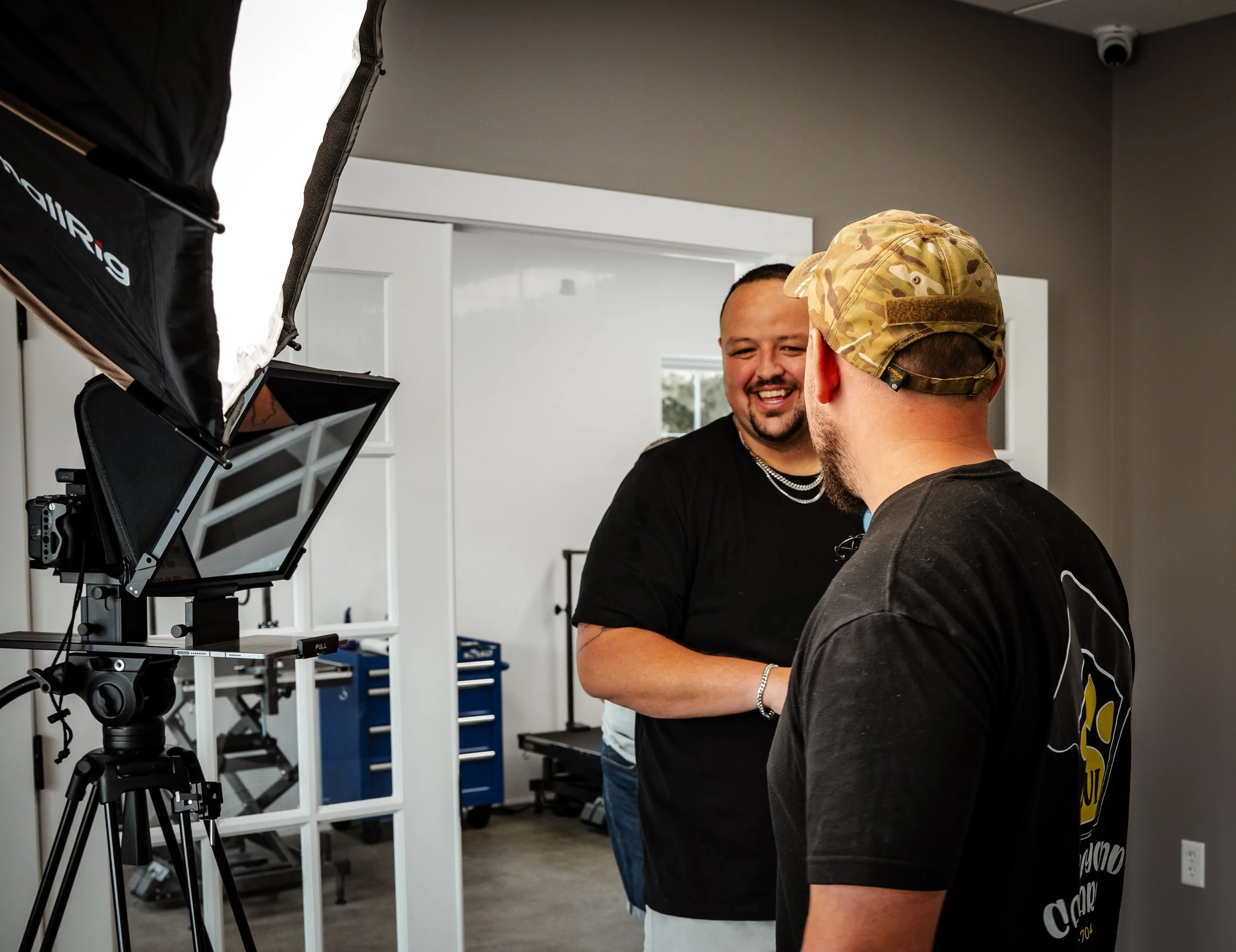 Two men on a photo shoot, one wearing a camouflage cap and the other smiling, with professional lighting equipment in a room with white walls and a window.