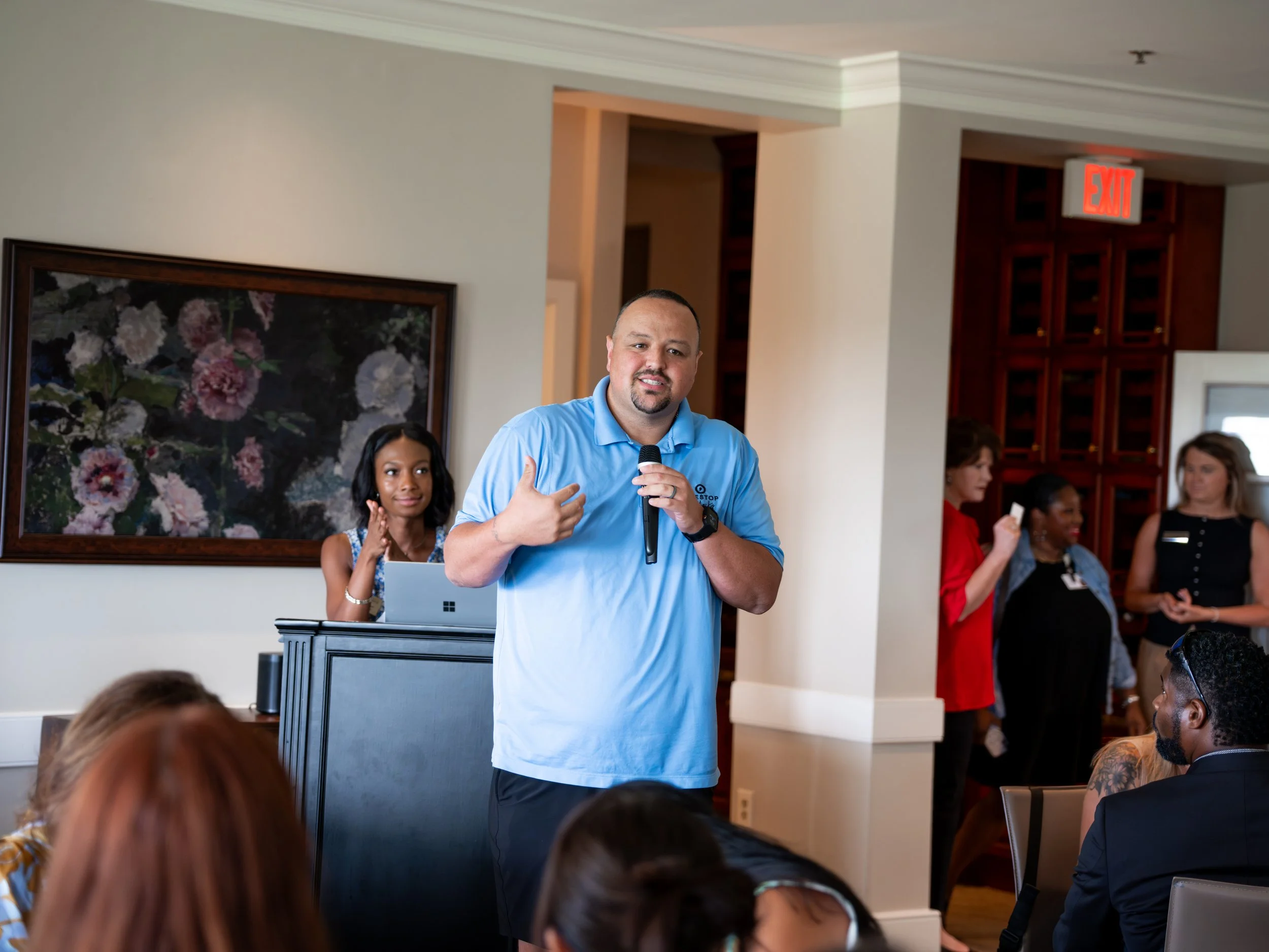 A man wearing a light blue polo shirt is speaking into a microphone while gesturing with his left hand. He is standing in front of a group of people in a room with light-colored walls, a Large floral painting on the wall, and an exit sign above a wooden door.
