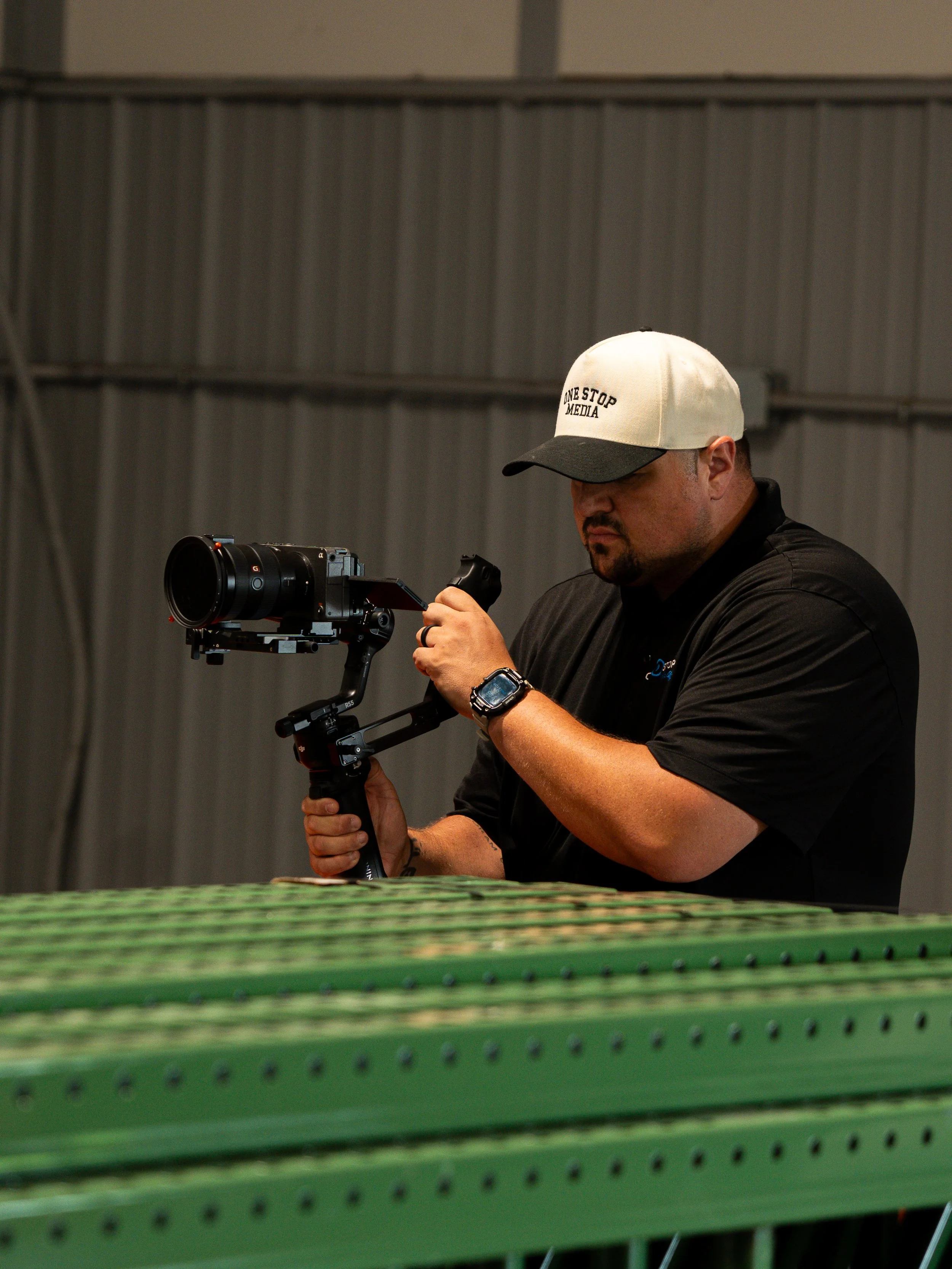 A man wearing a black shirt, a white and black cap, and a watch is operating a camera mounted on a stabilizer in an indoor setting with gray walls. The camera is pointed downward, and the man is looking at it while holding the stabilizer.