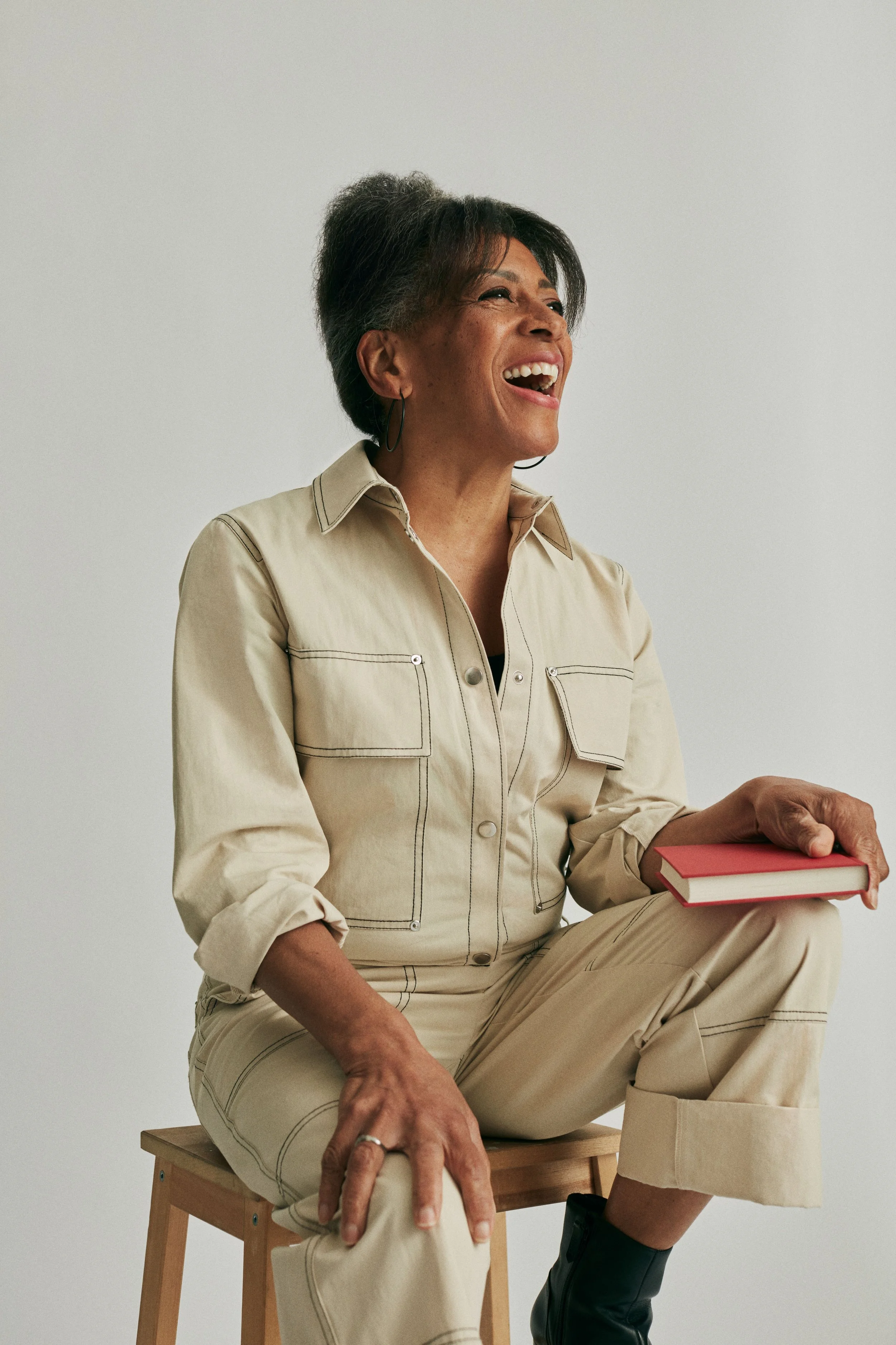 A woman with short gray hair sitting on a wooden stool, holding a red book, smiling and looking off to the side, against a plain light gray background.