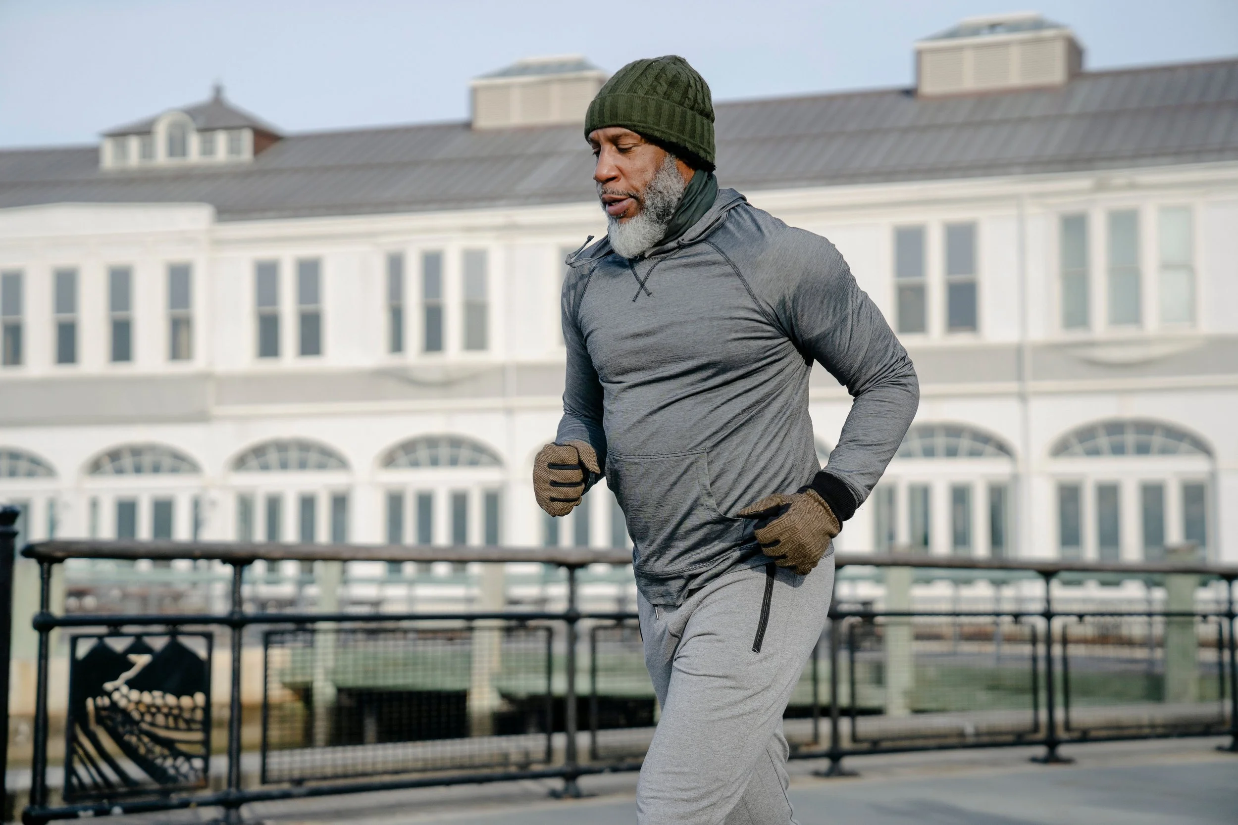 A man jogging outdoors in cold weather, wearing a green knit cap, gray athletic hoodie, gloves, and light gray sweatpants, with a building in the background.