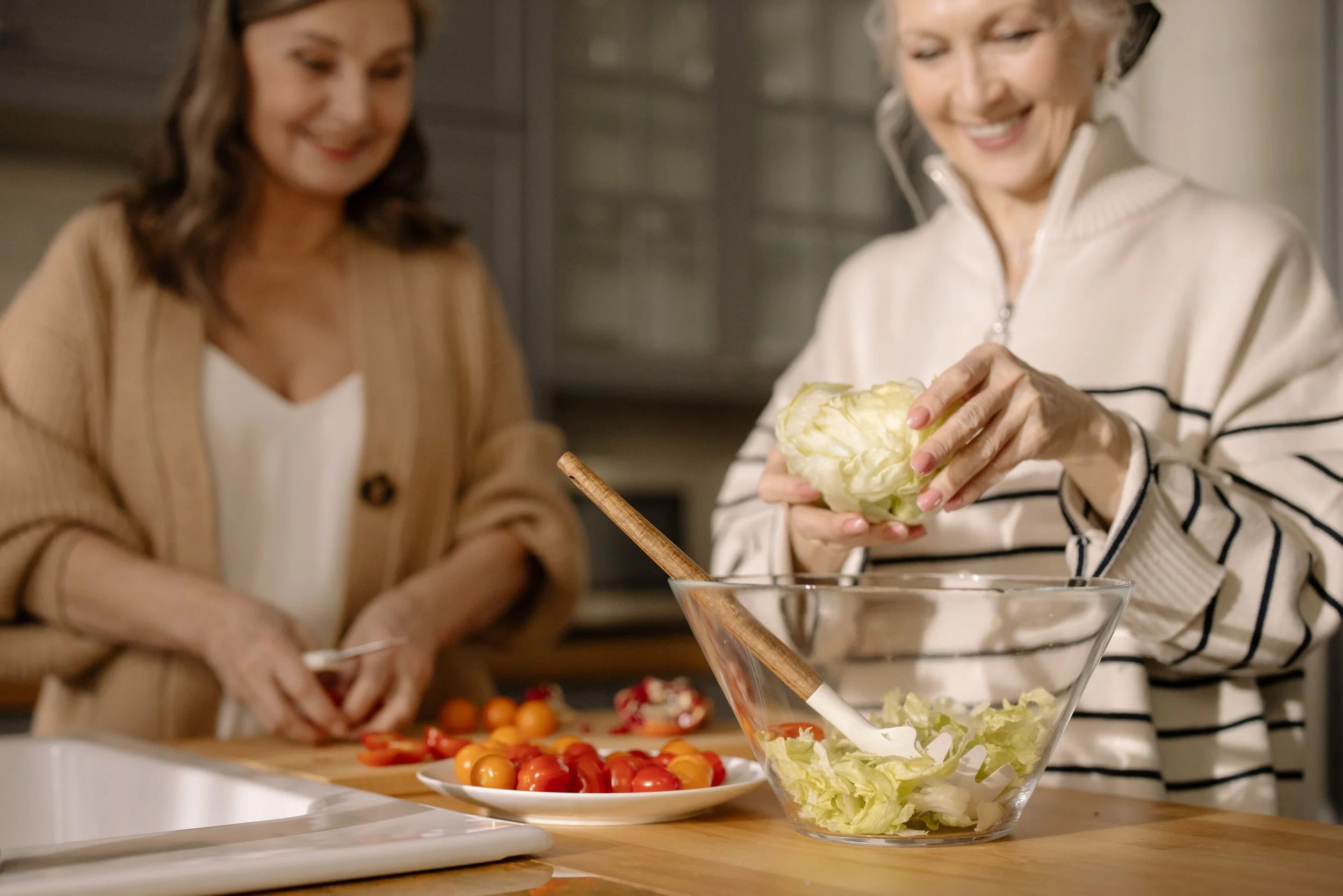 Two women preparing a salad in a kitchen, with cherry tomatoes on a plate and a bowl of lettuce on the counter.