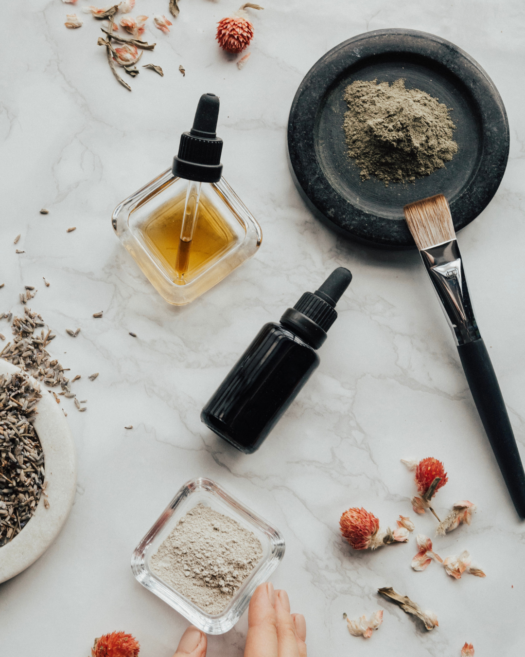 Spa or skincare setup with bottles of oil, a bowl of herbal powder, a makeup brush, dried flowers, and loose herbs on a marble surface.