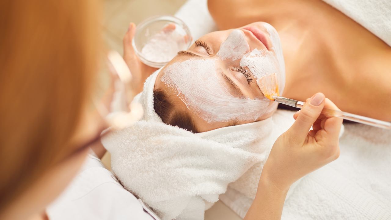 A woman receiving a facial treatment, lying down with her eyes closed, as an esthetician applies a white face mask with a brush.
