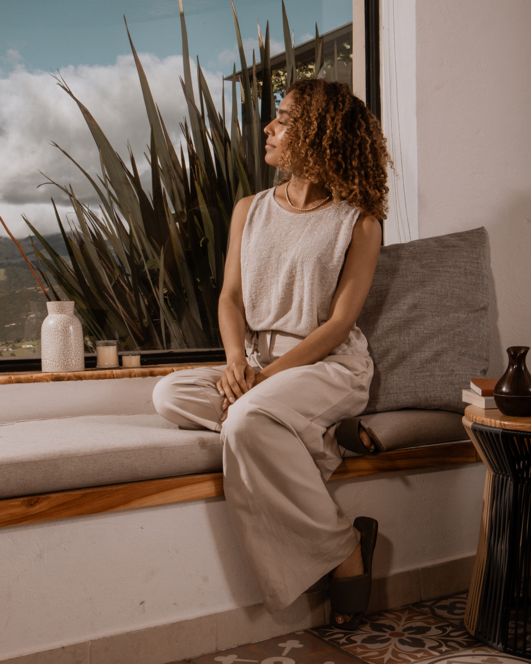 Woman sitting on a window seat with a large plant behind her, looking out the window with eyes closed. Candles and a decorative vase are on the windowsill, and a woven table with books and a dark pitcher are nearby.