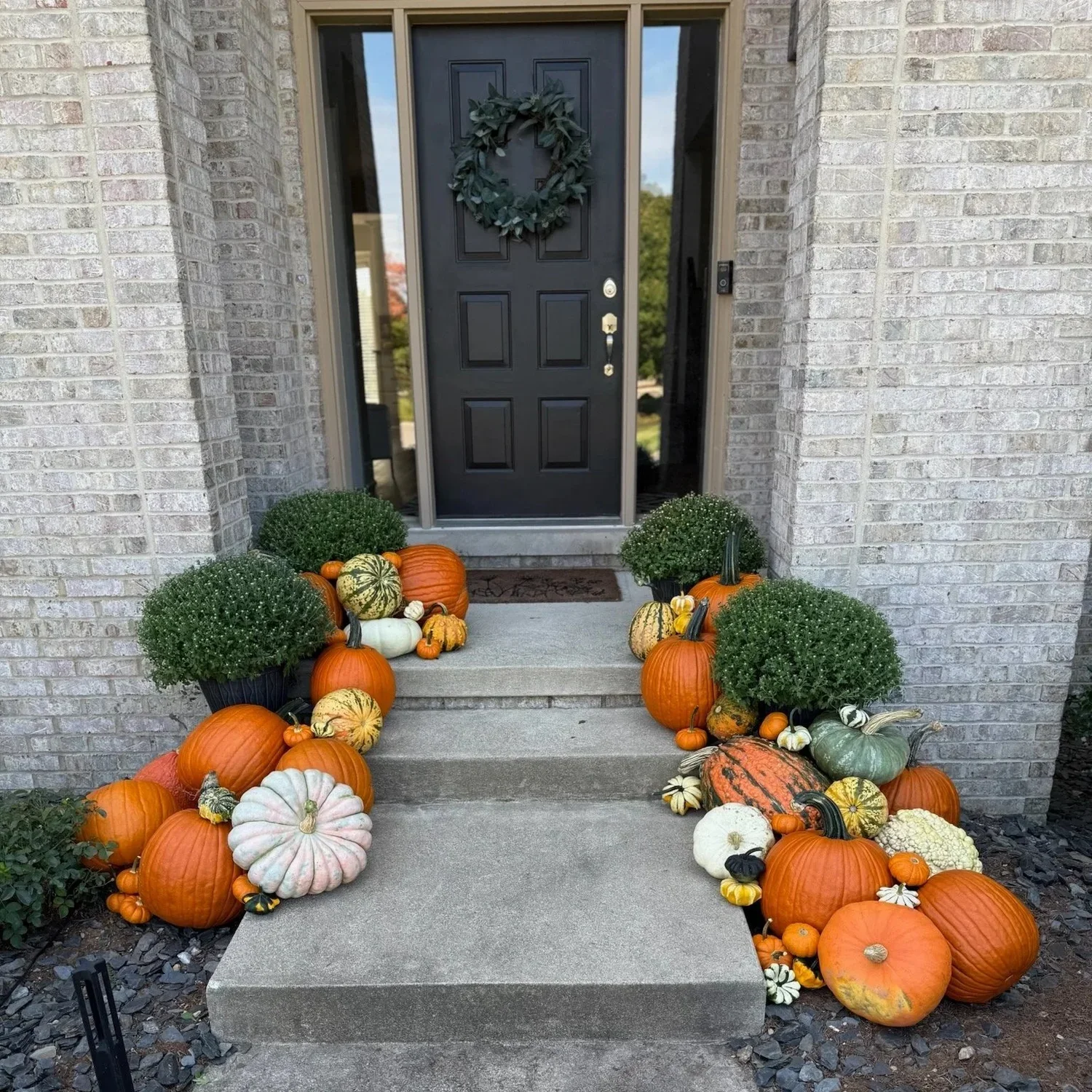 Front porch decorated with various pumpkins and gourds, flanked by potted plants and situated in front of a brick house with a black door adorned with a green wreath.