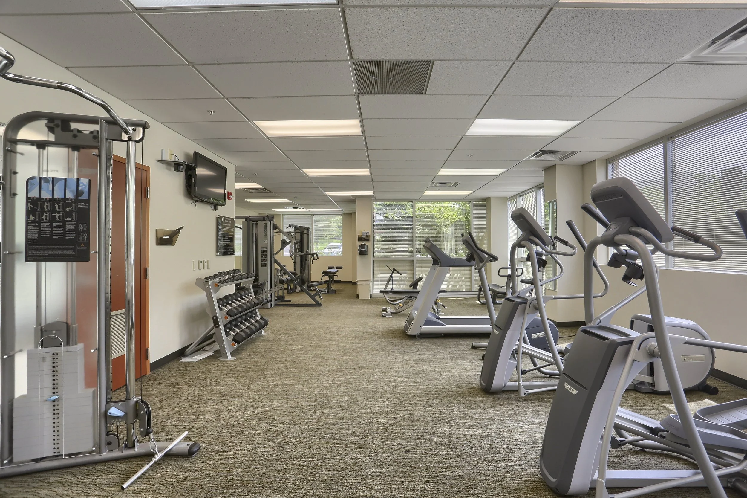 Fitness room with cardio equipment, weight machines, a flat-screen TV, and large windows with blinds.