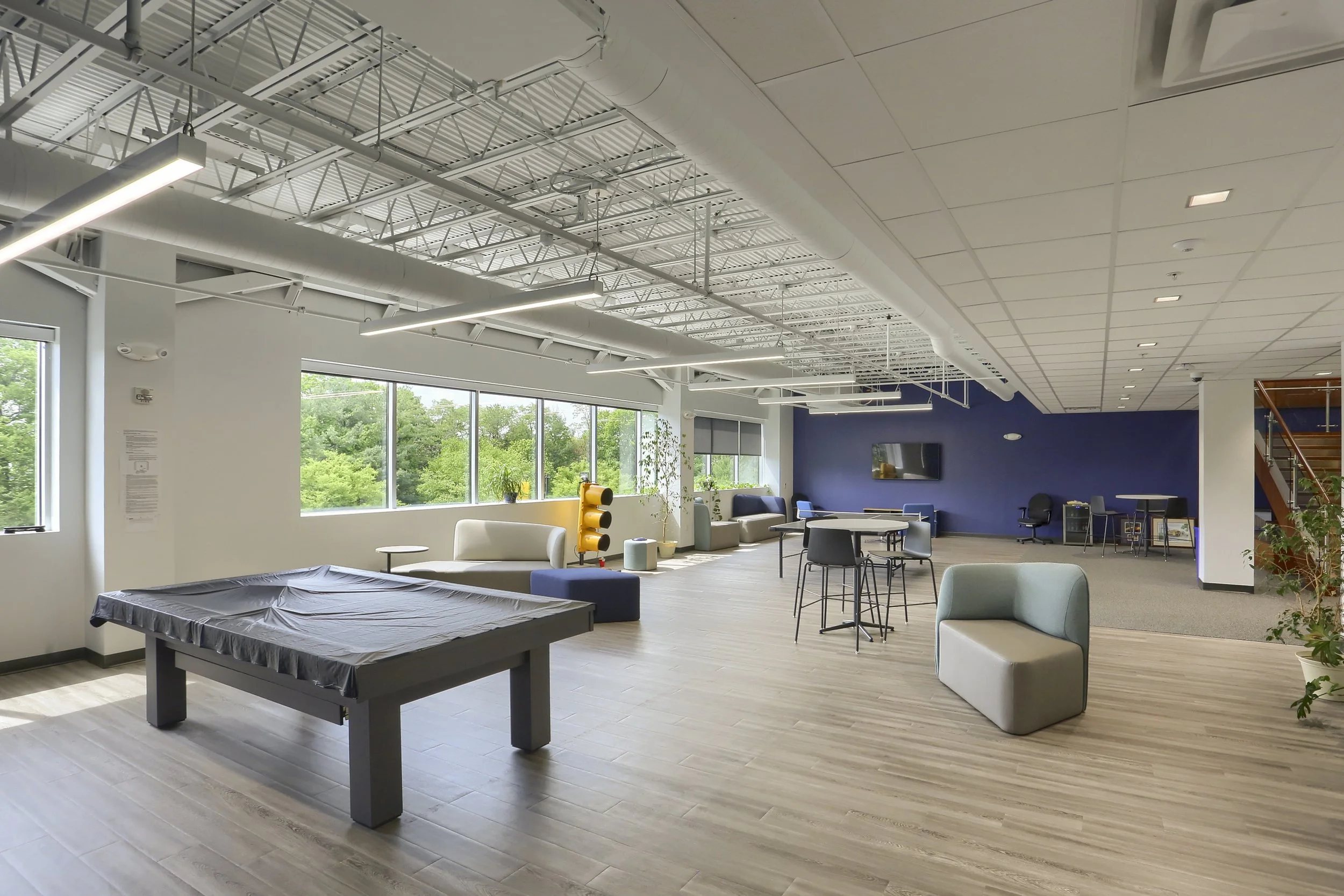 Modern lounge area with large windows, white and navy walls, and various seating options including sofas, armchairs, and high tables with stools. Overhead, there are bright linear lights and exposed ceiling beams with air ducts, in a spacious and well-lit environment.