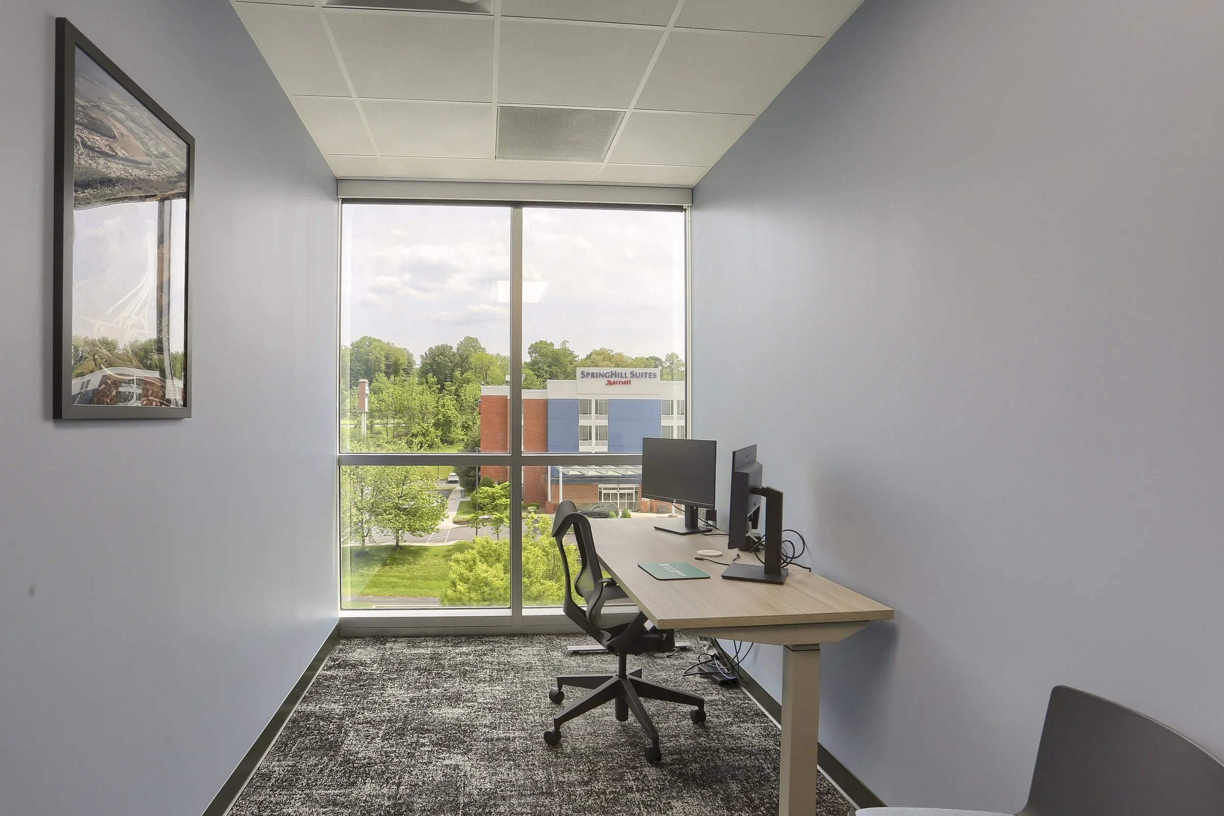 Office workspace with a desk, office chair, computer monitors, and a large window showing trees and a building with a sign for SpringHill Suites Marriott.