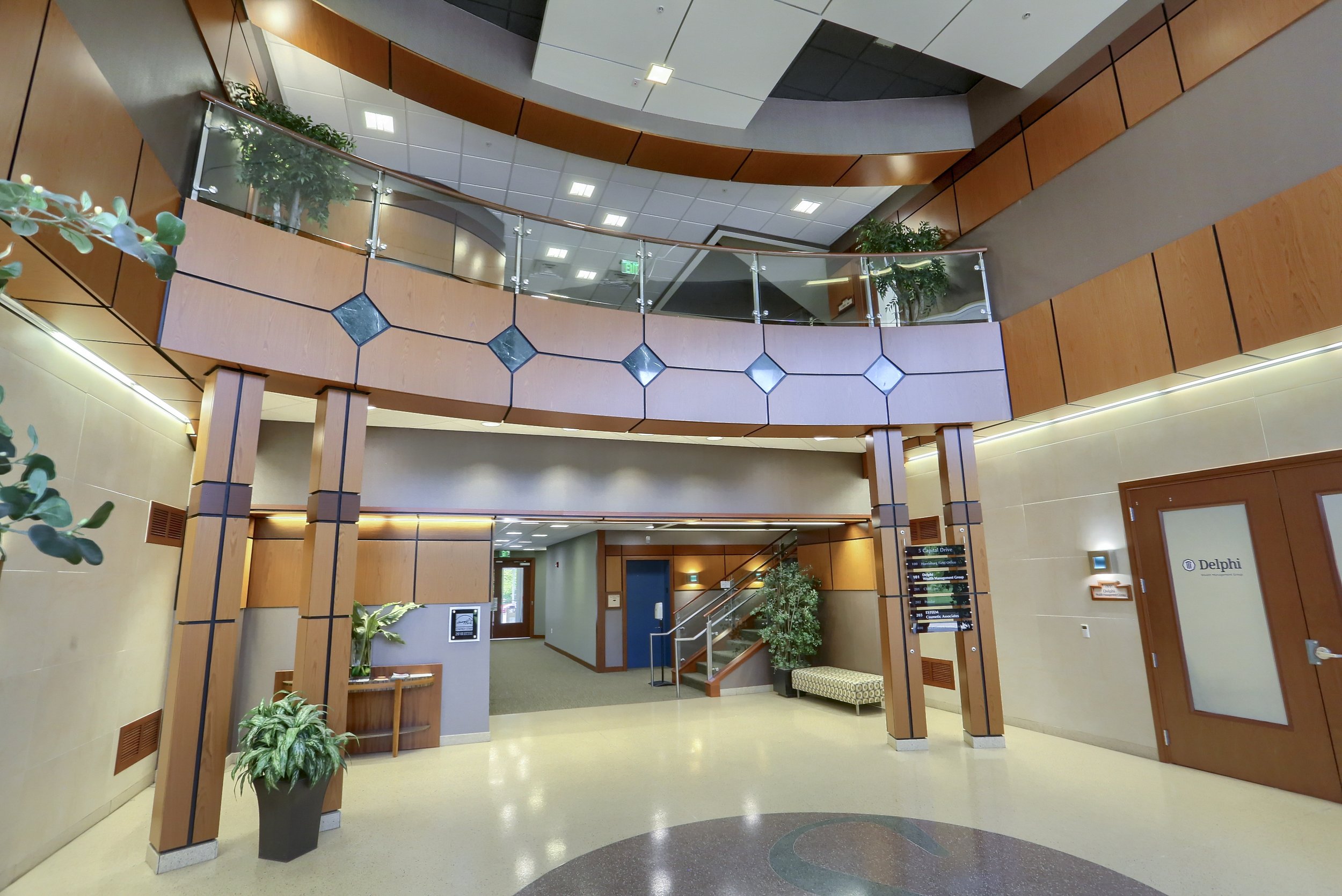 Interior of a modern office building lobby with wood-paneled walls, potted plants, a staircase, and directional signs on the wall.