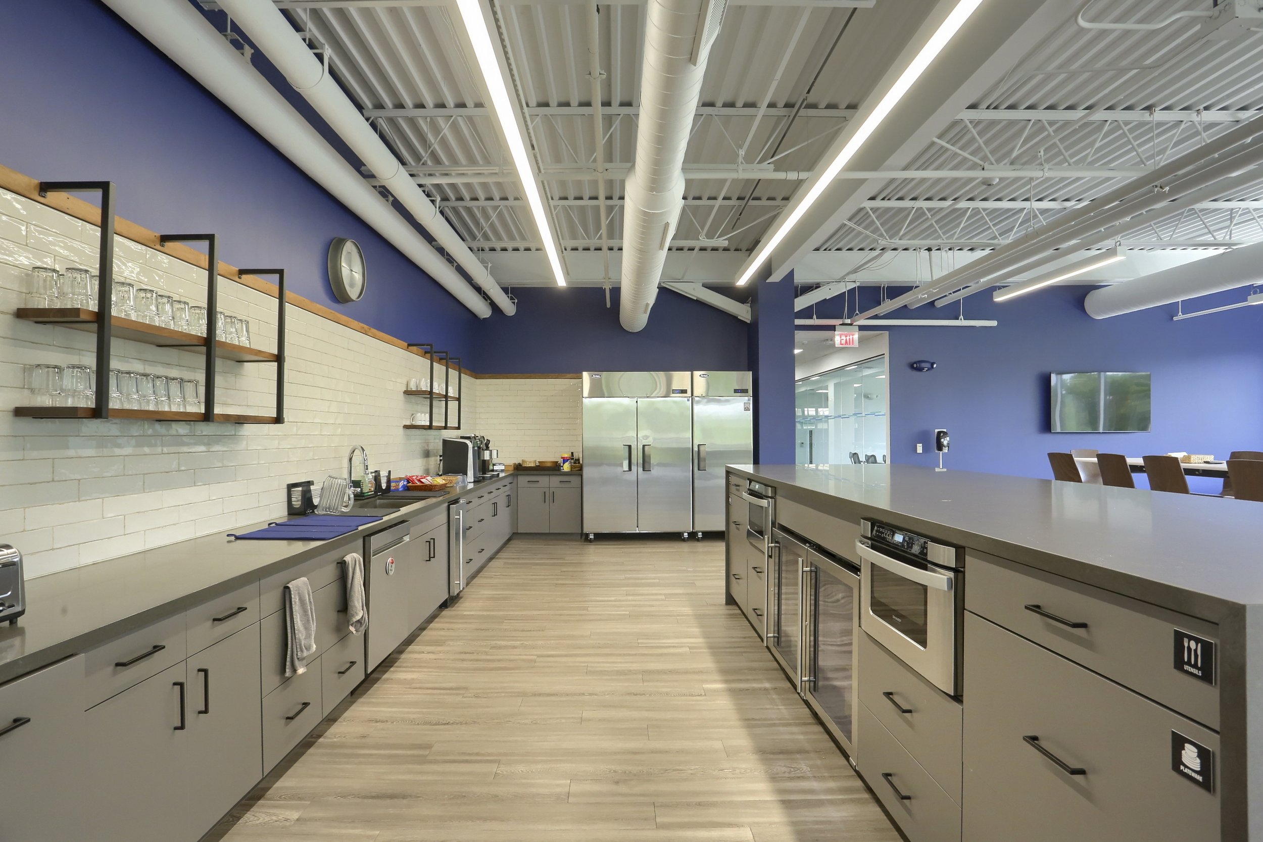 Modern kitchen with gray cabinets, stainless steel appliances, open shelving with glasses, white brick backsplash, blue accent wall, ceiling with exposed ductwork and linear lighting, wood flooring, TV on wall, and a view into an adjacent room.