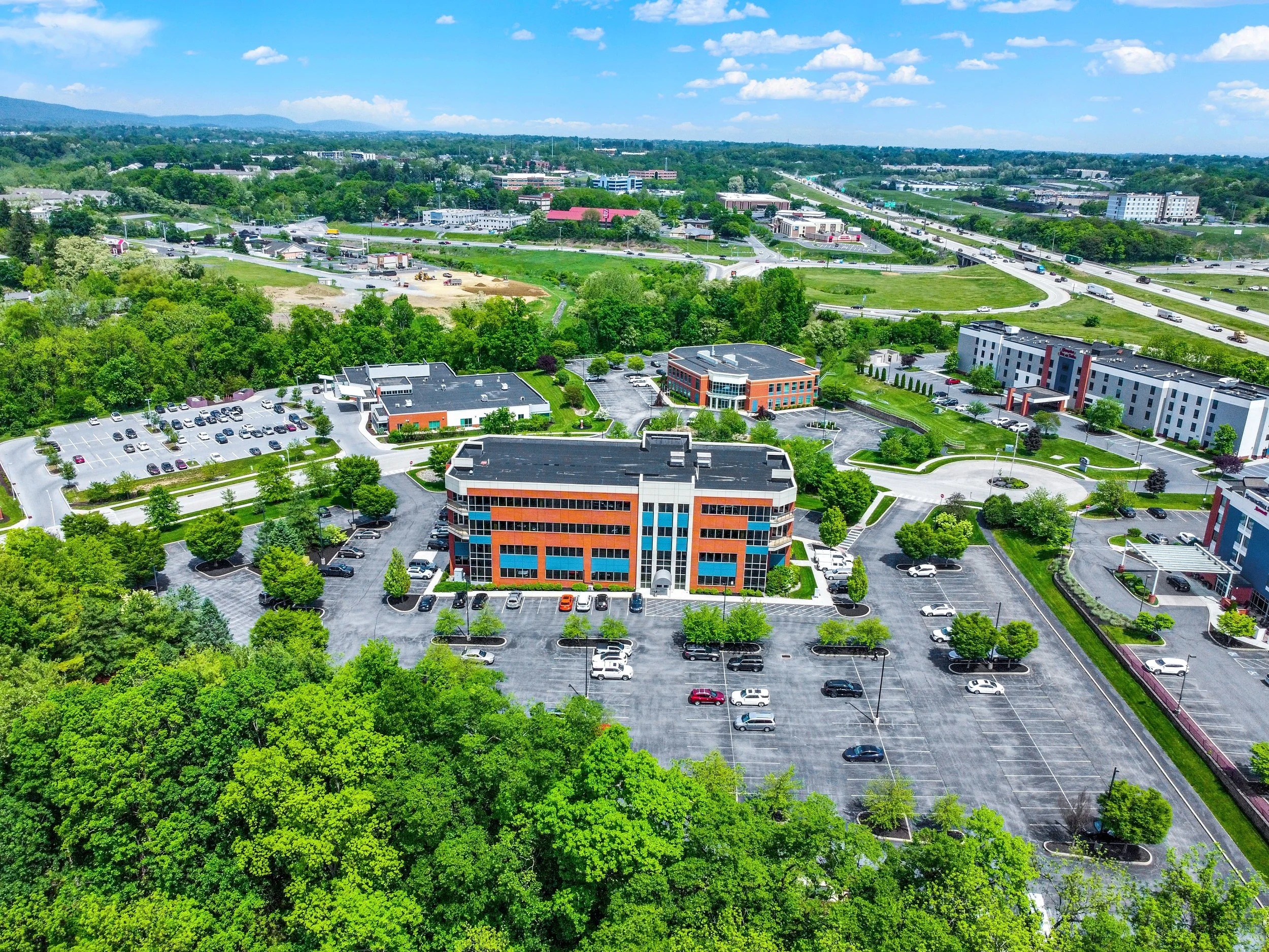 An aerial view of a cityscape with buildings, parking lots, roads, and greenery under a partly cloudy sky.