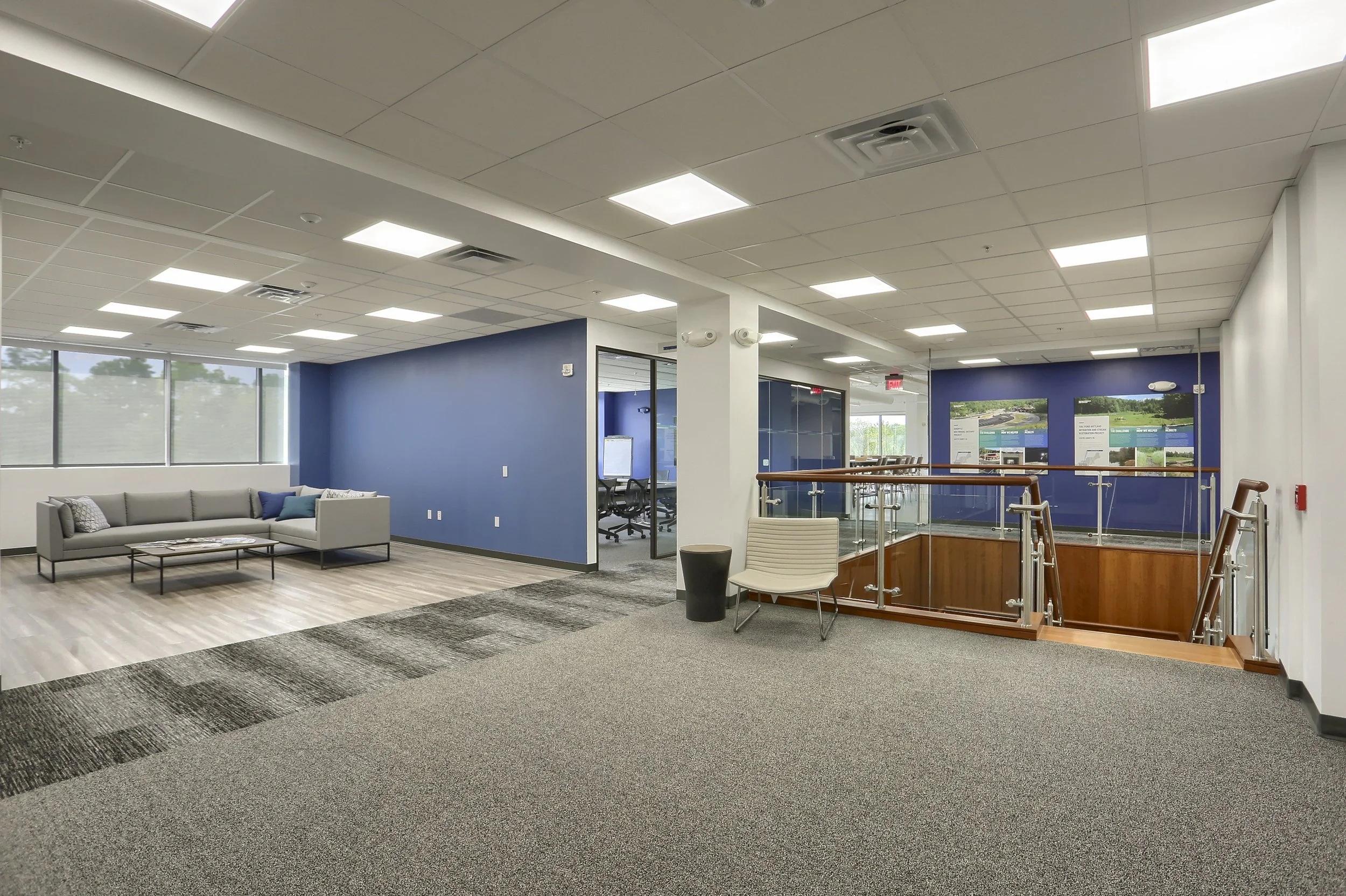 A modern office lobby area with a sitting area featuring a gray sofa and a black coffee table, large windows letting in natural light, blue accent walls, a staircase with a glass railing, informational display boards on the wall, and a conference room in the background.