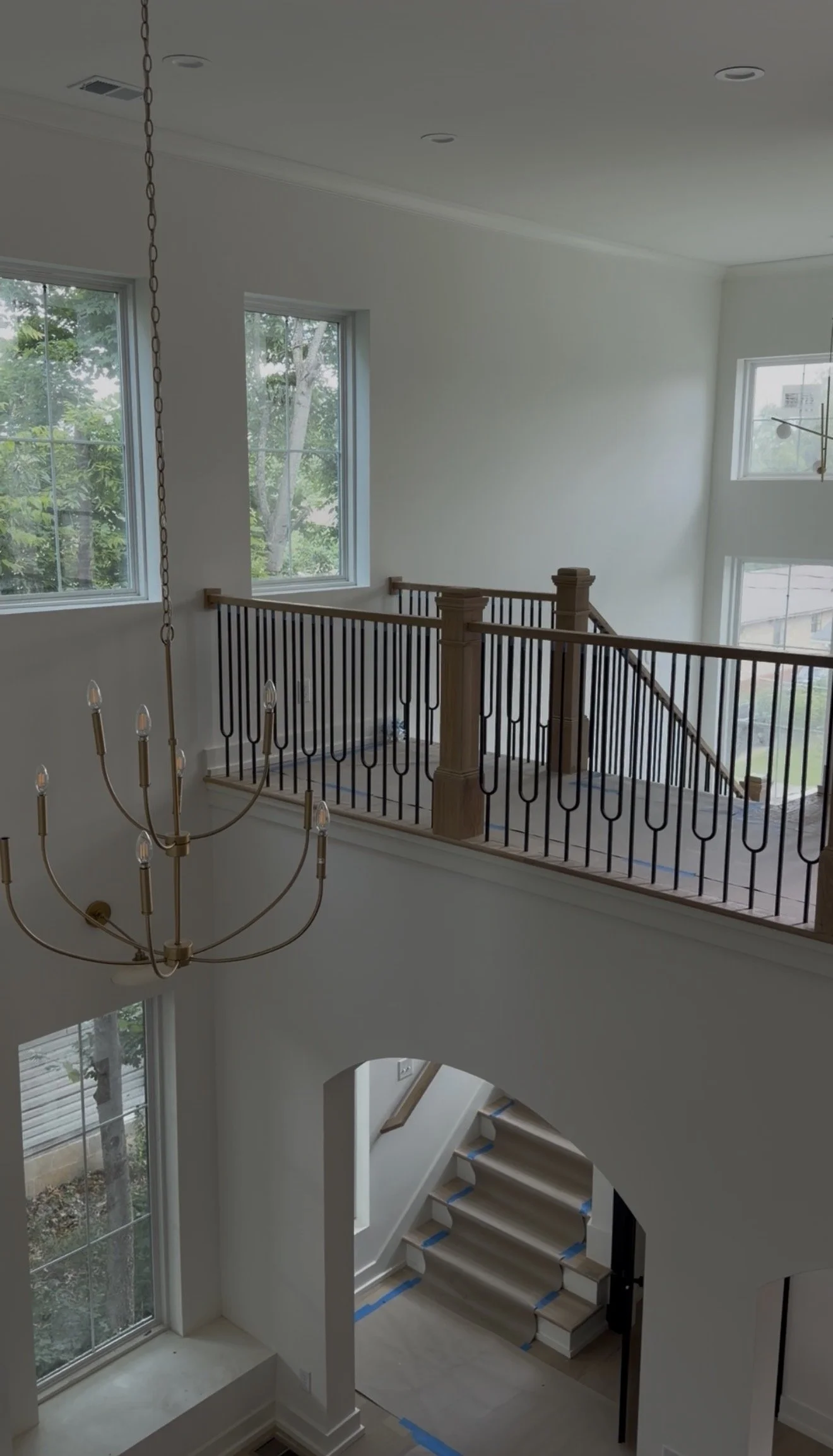 Interior view of a house under construction, showing a staircase with beige steps covered with protective tape, a wooden and black metal railing at the second floor, large windows letting in natural light, and a chandelier hanging from the ceiling.